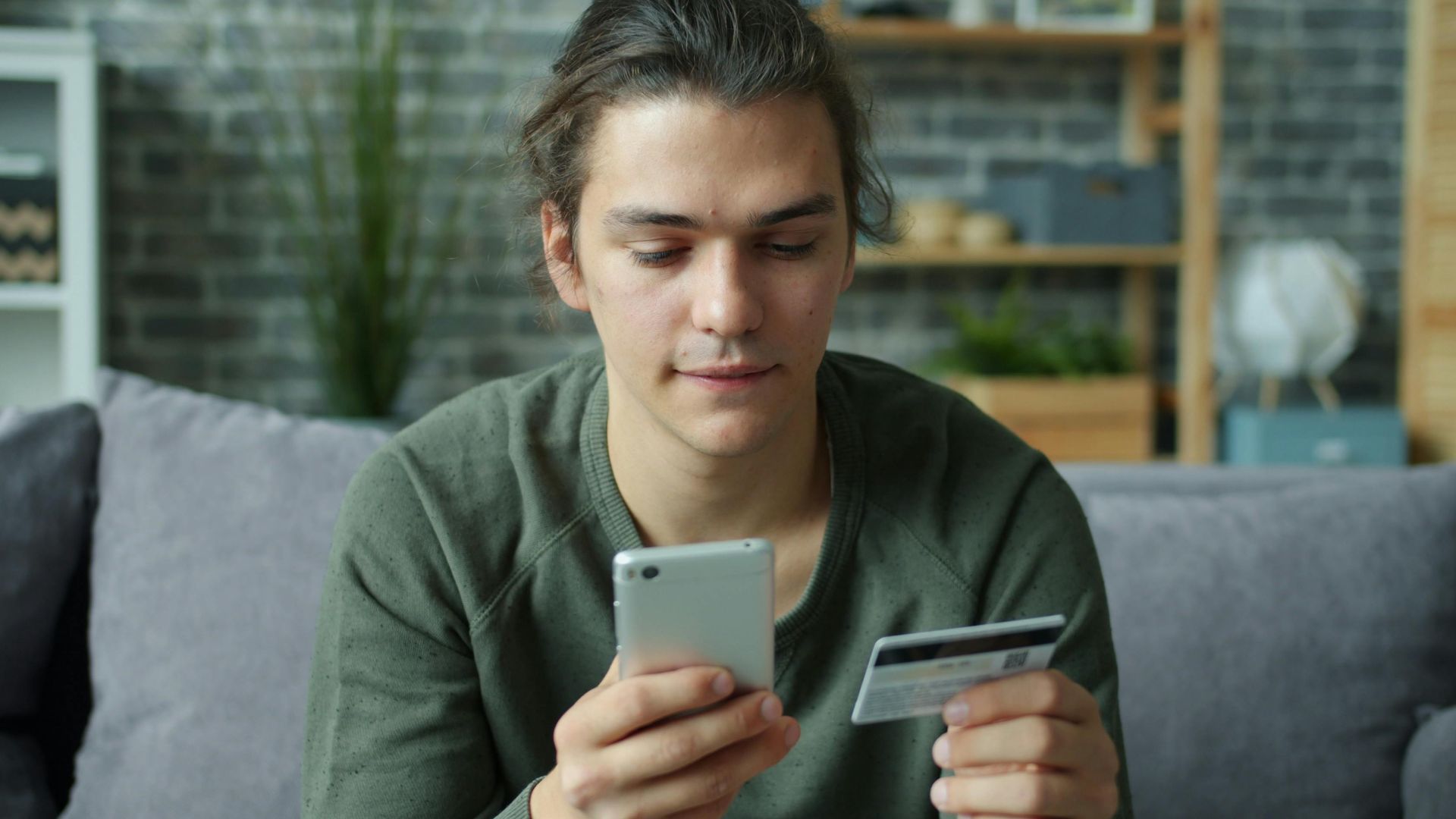 A young man using a smartphone and credit card for online shopping at home.