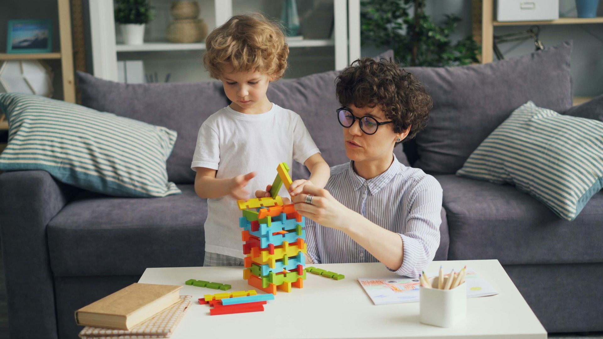 A mother and son playing with colorful building blocks on a table in a cozy living room.
