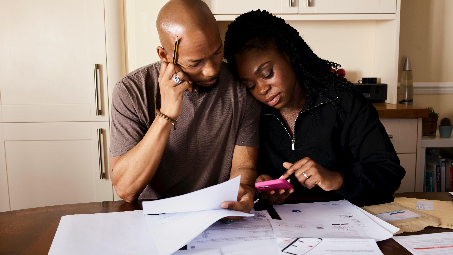 A couple sits at a table managing domestic finances, evaluating documents and using a smartphone.