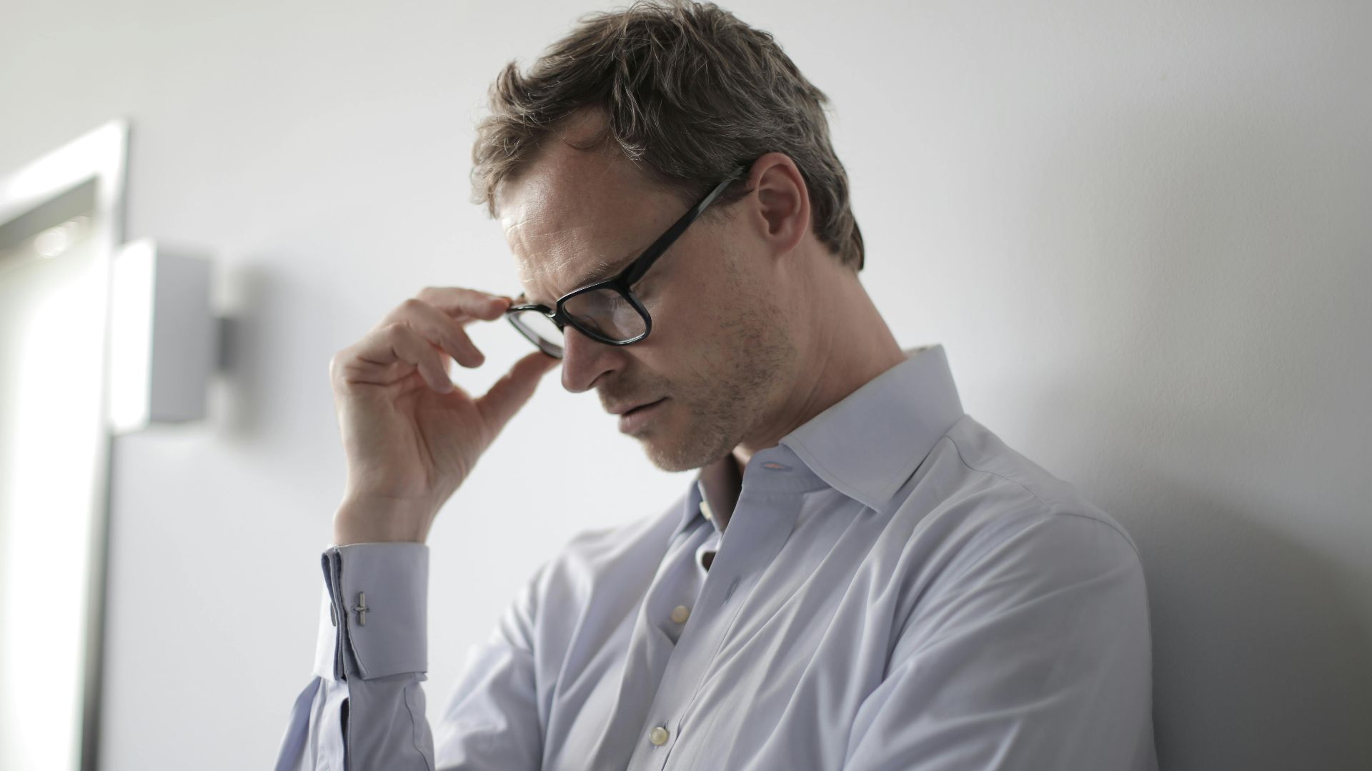 An adult man in a shirt and glasses appears stressed while leaning against a wall indoors.