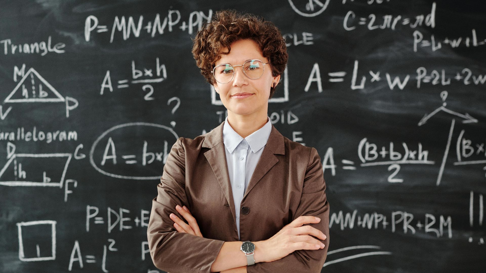 Confident female teacher standing with arms crossed in front of a detailed mathematical blackboard.