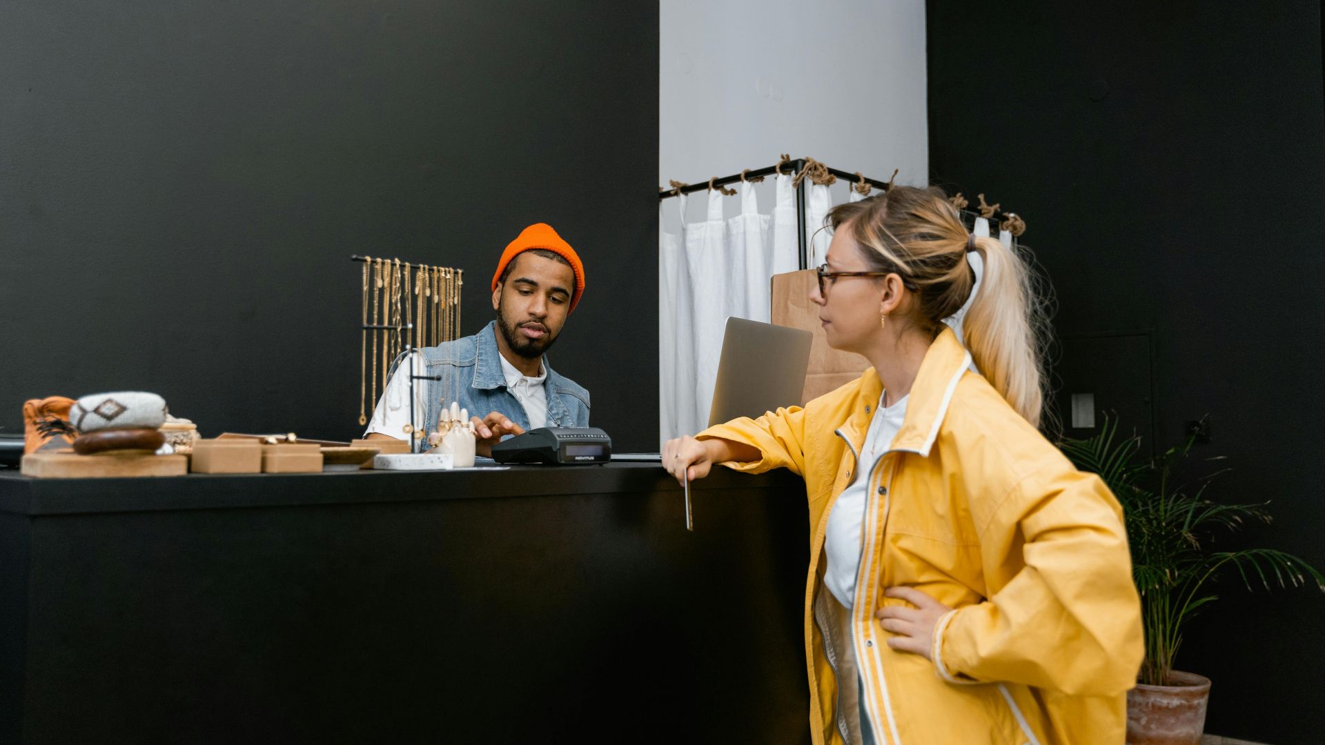 A woman pays with a card at a store counter to a bearded cashier in a denim vest.