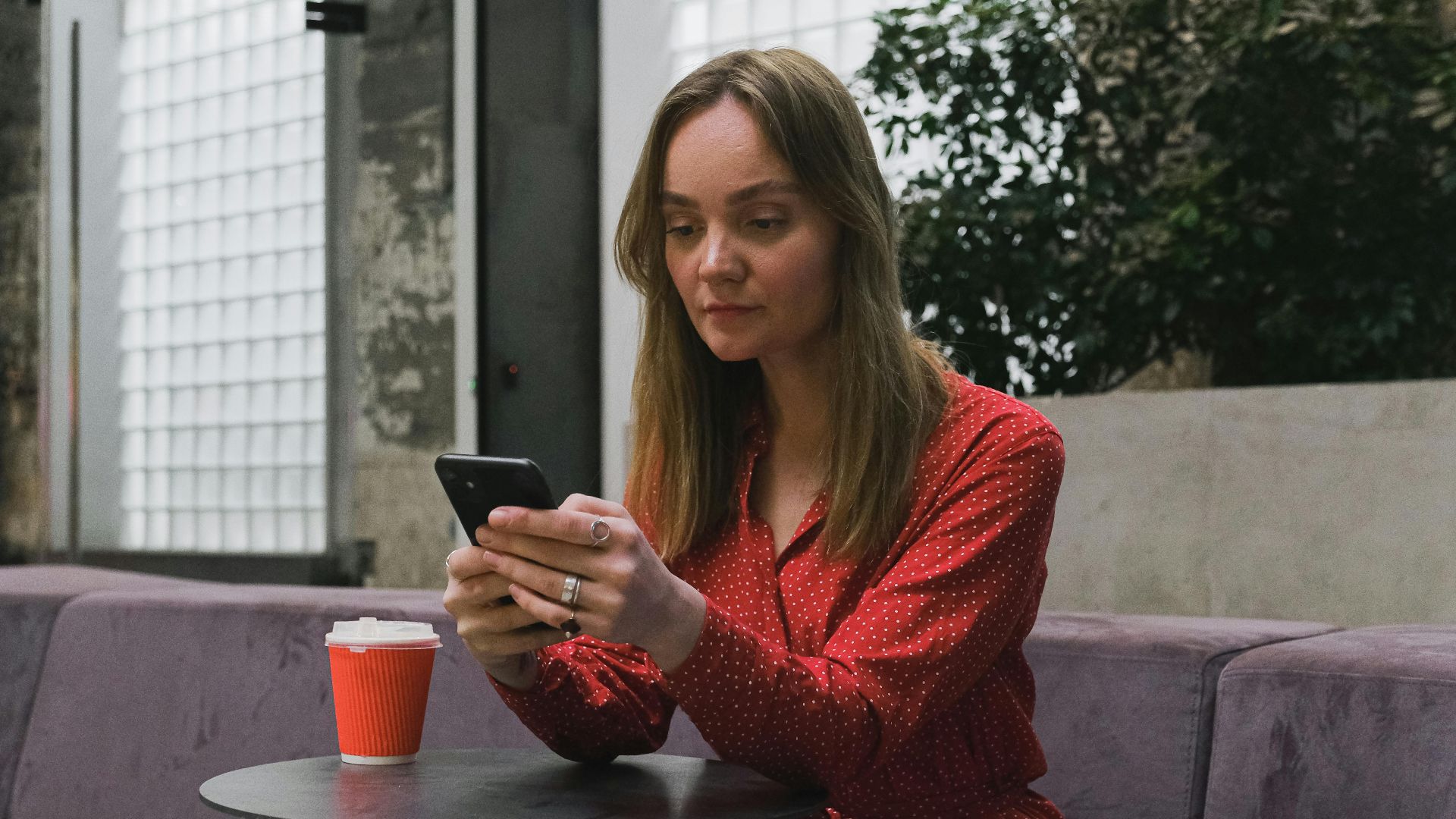 Young woman in red dress sitting indoors, using smartphone at a table with a cup of coffee.