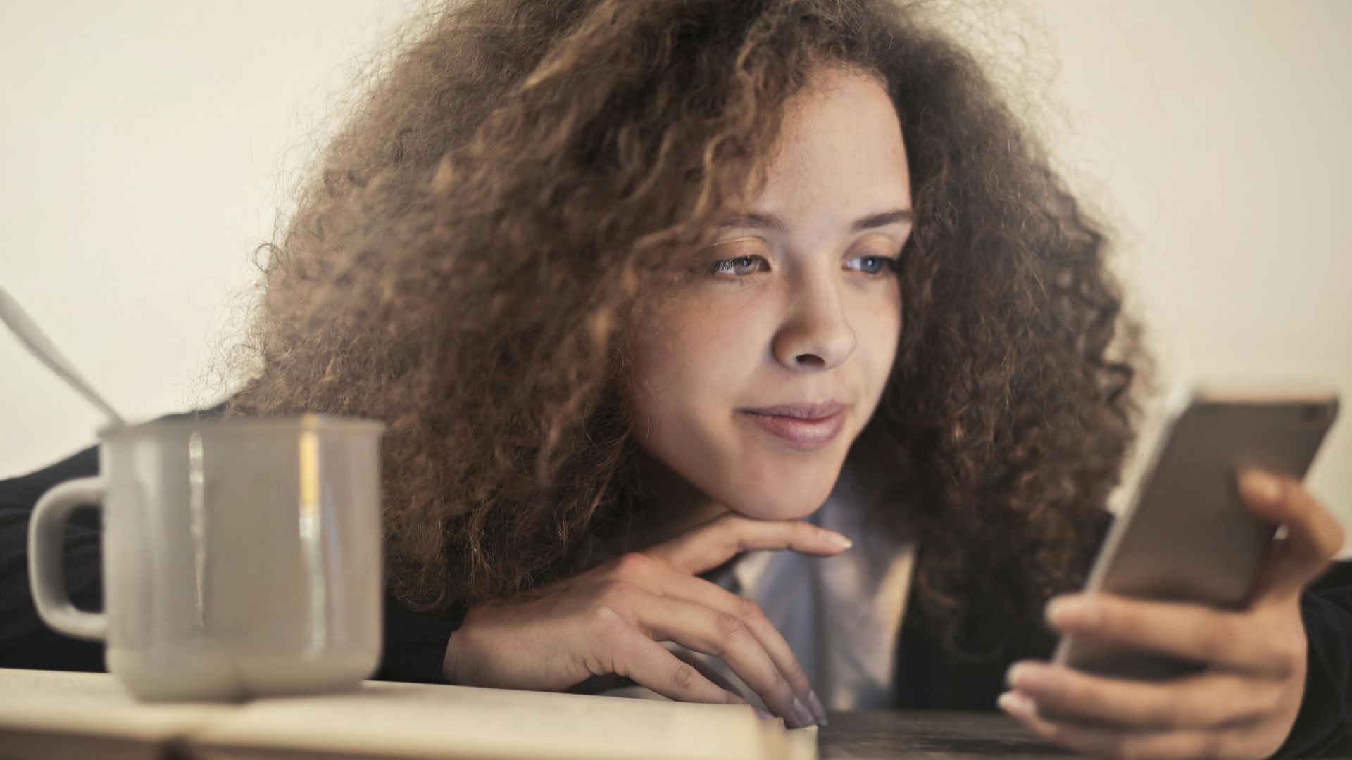 Young woman enjoying a relaxing moment while checking her smartphone indoors with a cup of coffee.
