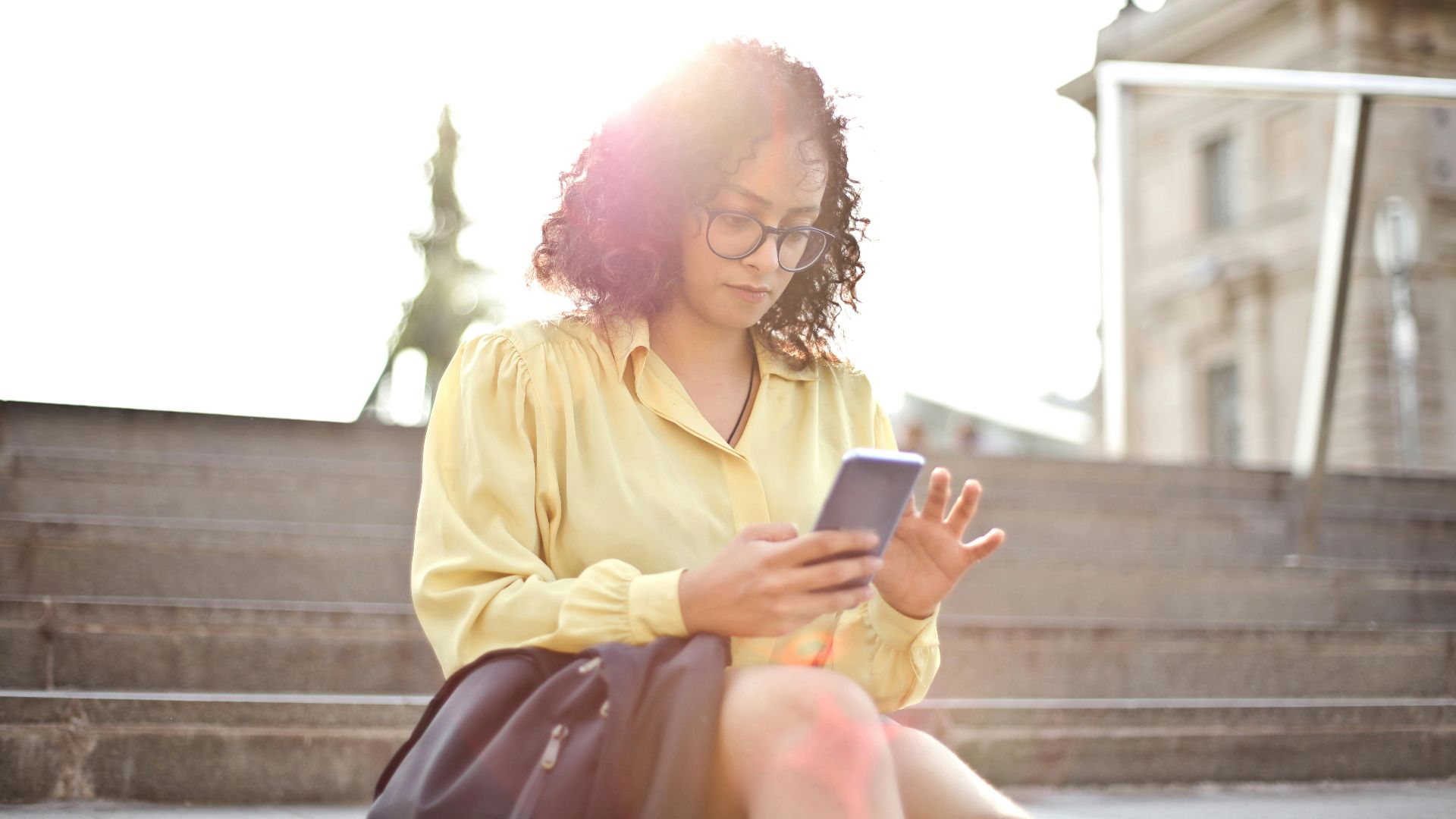 Young woman with curly hair and glasses texting on her smartphone while sitting on concrete stairs in daylight.