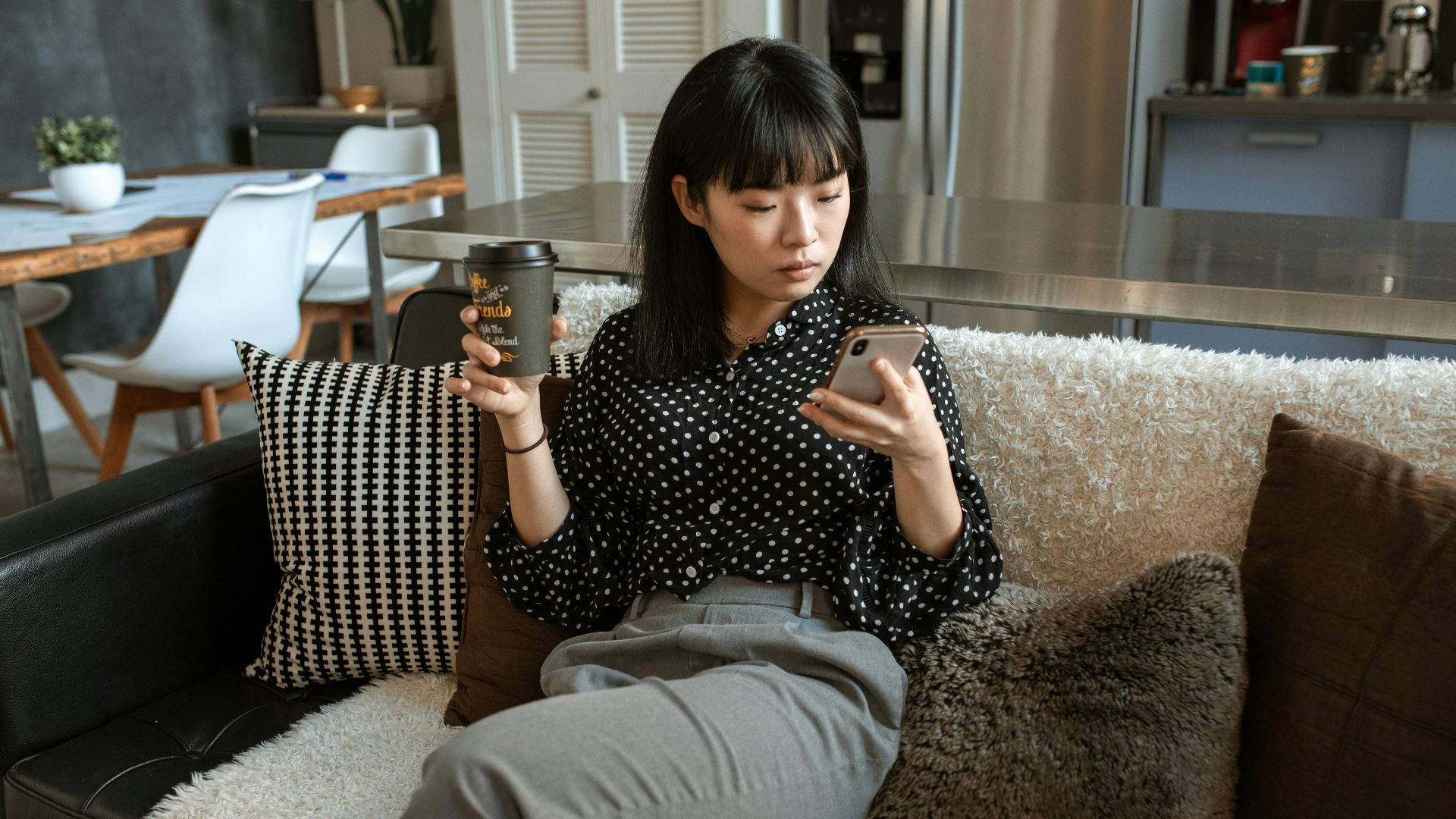 A young woman in a polka dot shirt enjoys coffee while using a smartphone in a cozy living room.