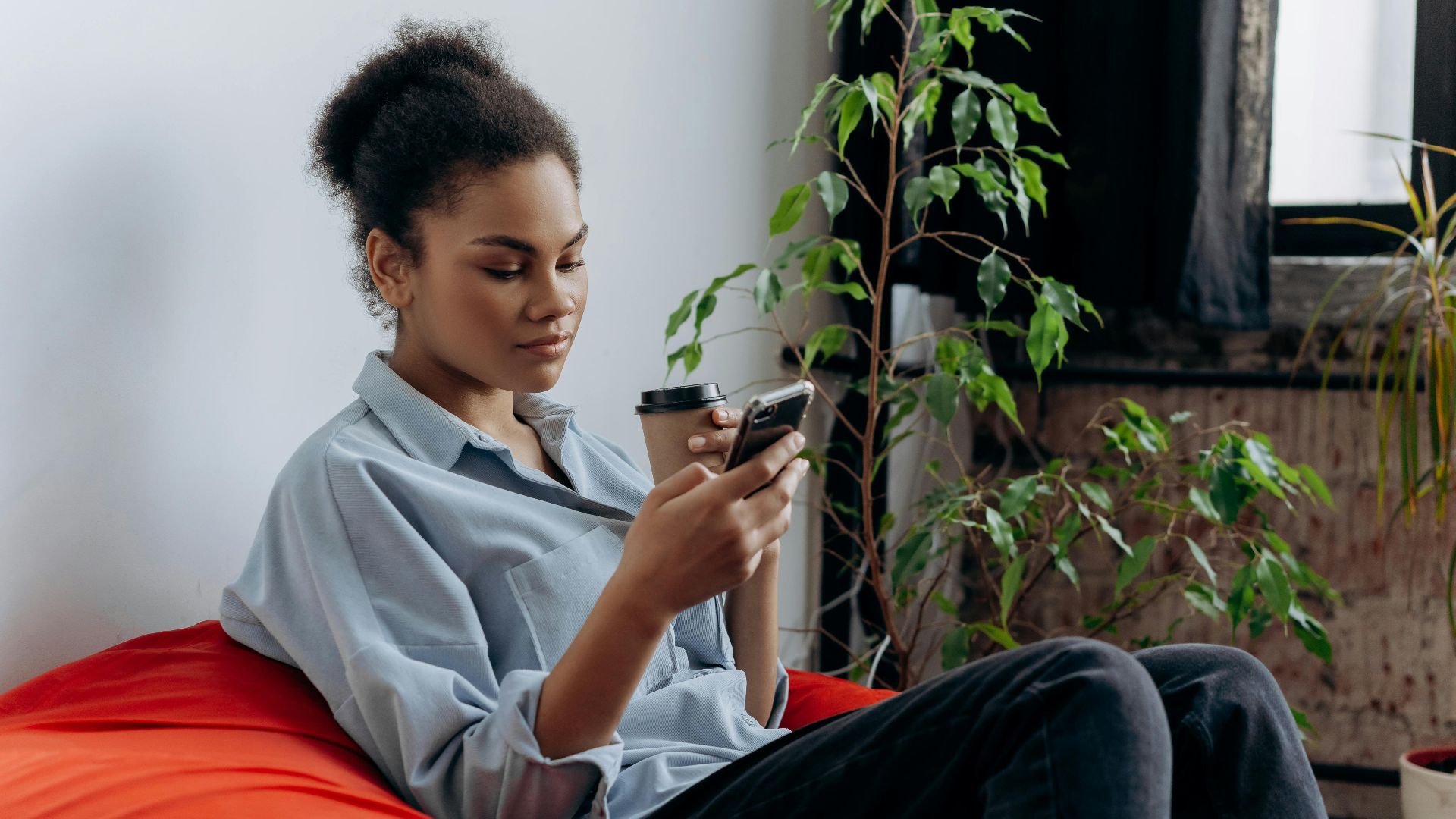 A young woman sits indoors, using her smartphone while sipping coffee, surrounded by plants.