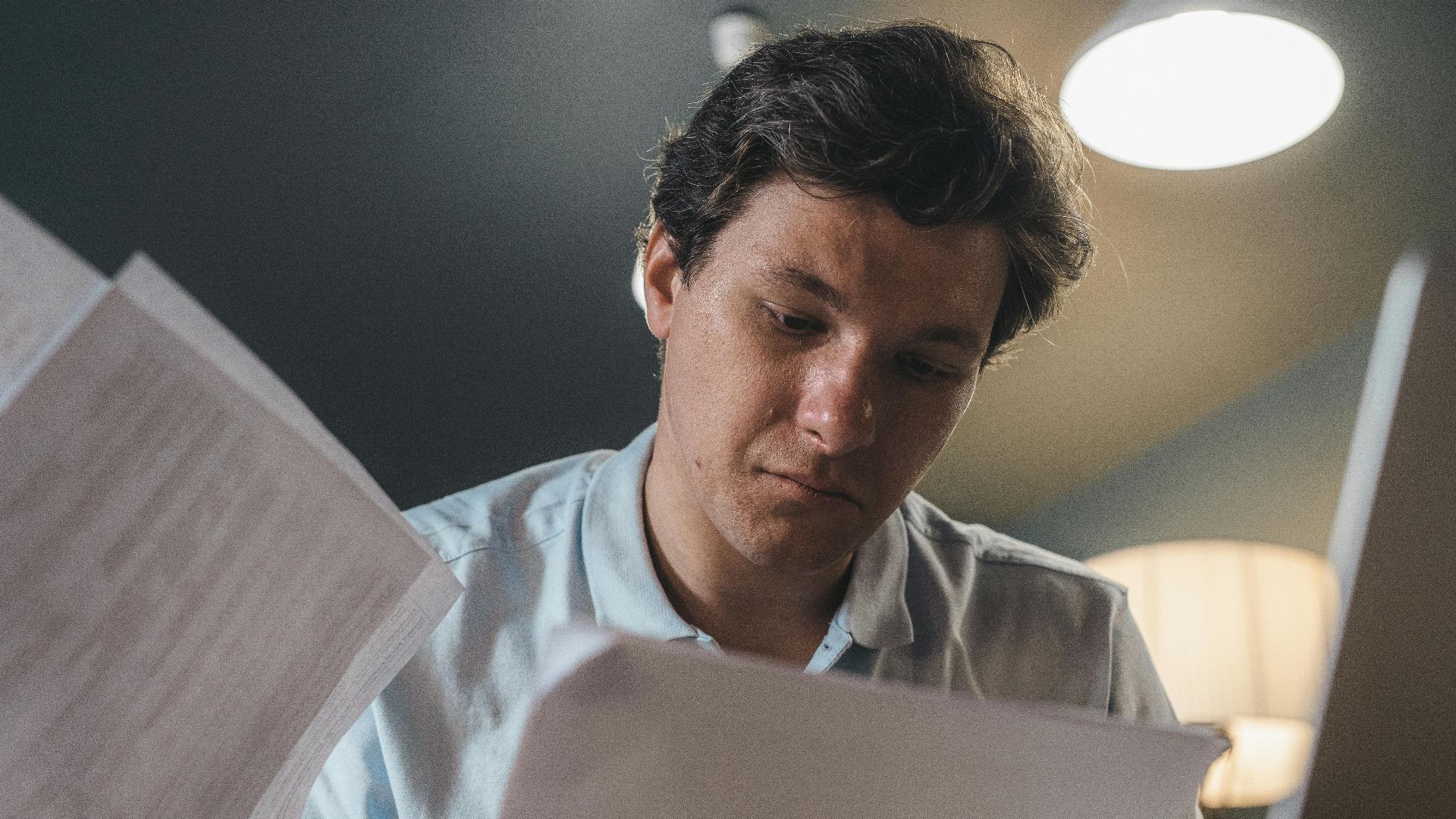 Serious man wearing a light blue shirt intently reading printed papers indoors under warm lighting.
