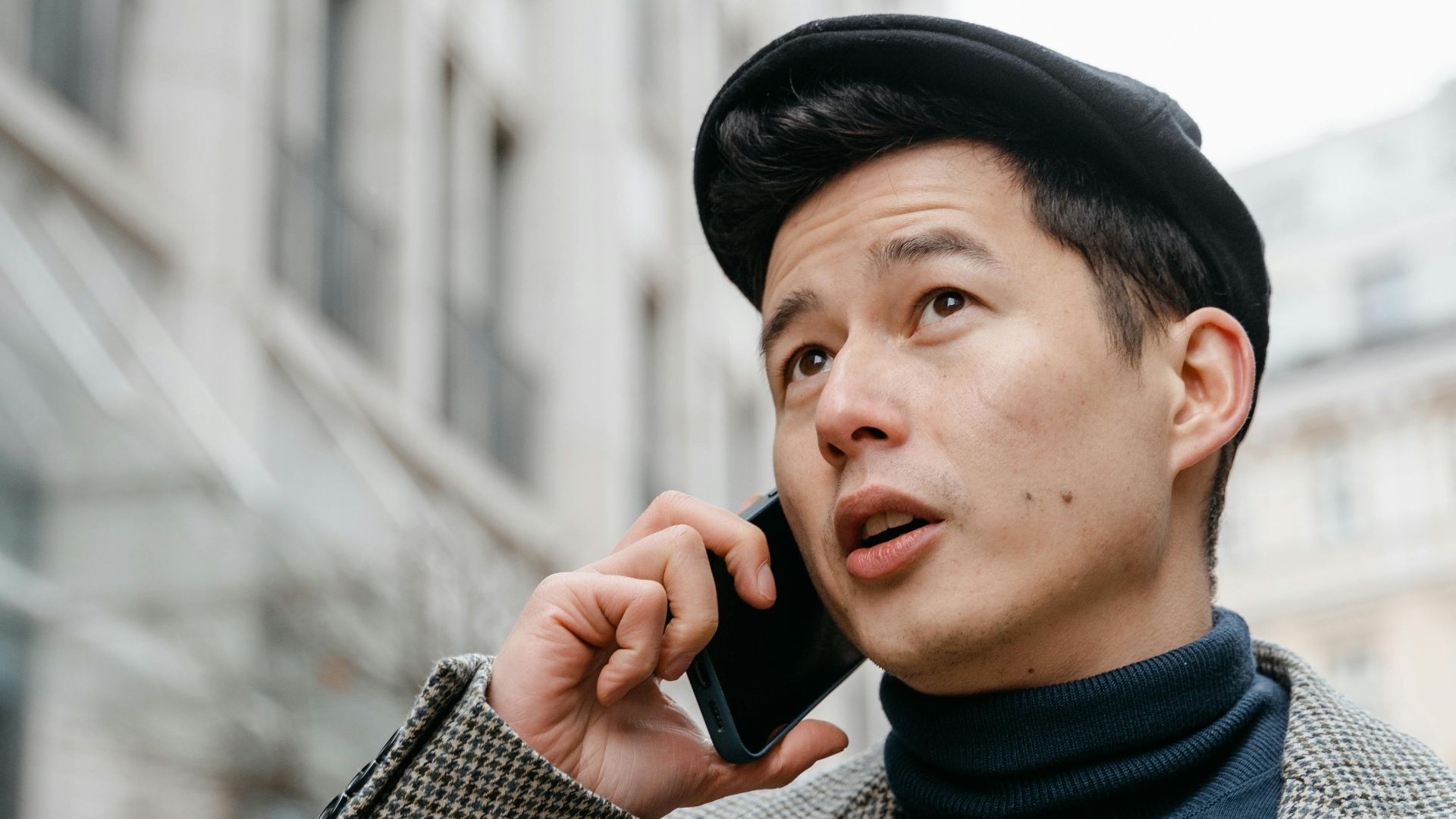 Stylish man in a city talking on a smartphone outdoors, wearing a coat and hat.