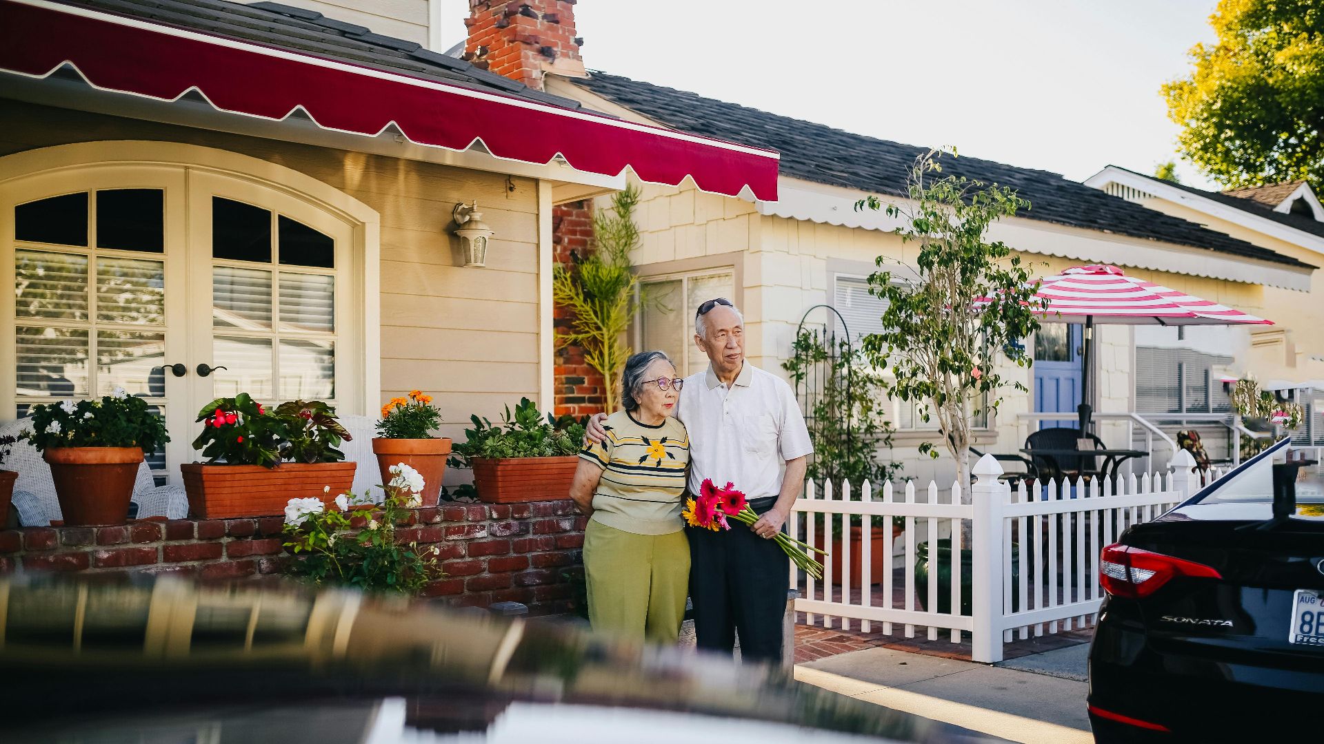 An elderly couple stands in front of a charming house holding a bouquet, capturing a serene lifestyle moment.