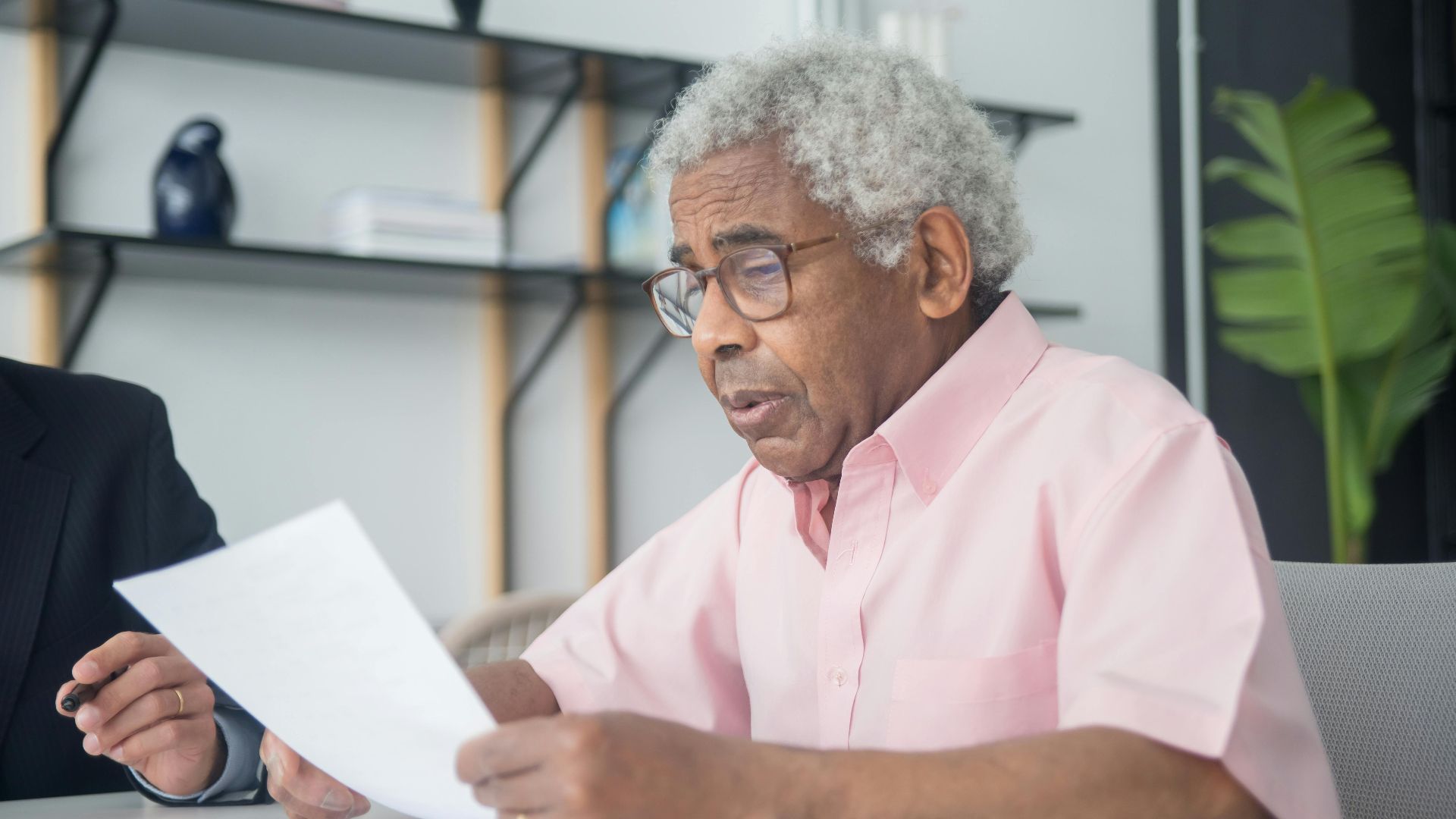 Elderly man with glasses reading a document in a home office setting, reflecting focus and concentration.