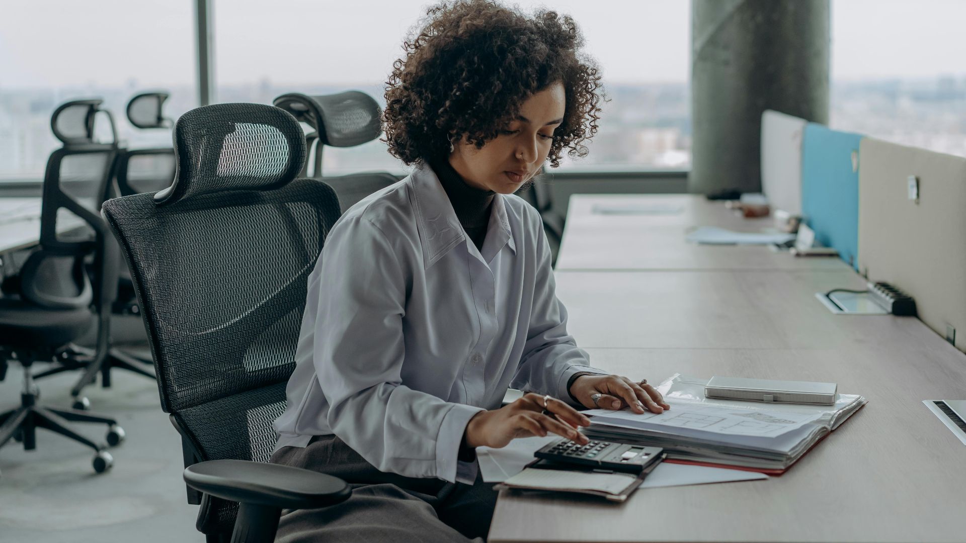 A professional woman with curly hair calculating documents in a modern office setting.