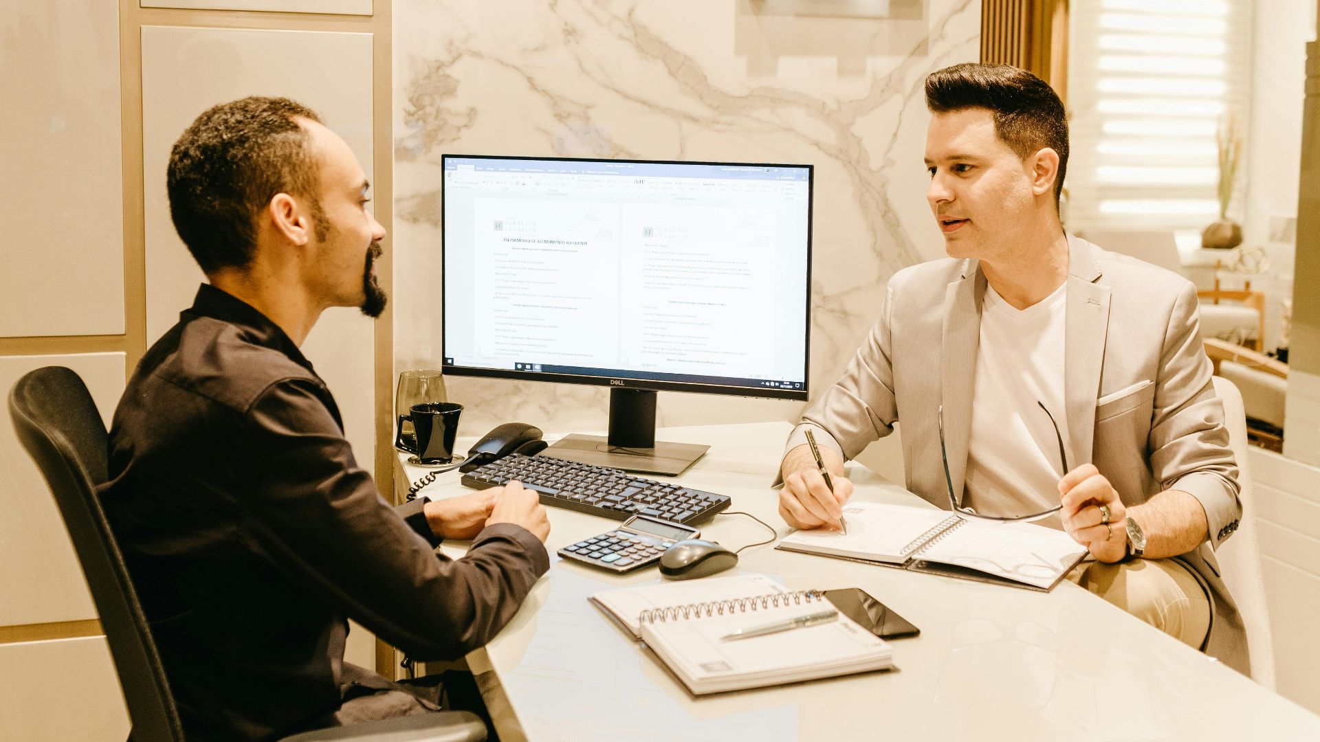 Two businessmen discussing work at a sleek and modern office desk, using computer and notes for collaboration.