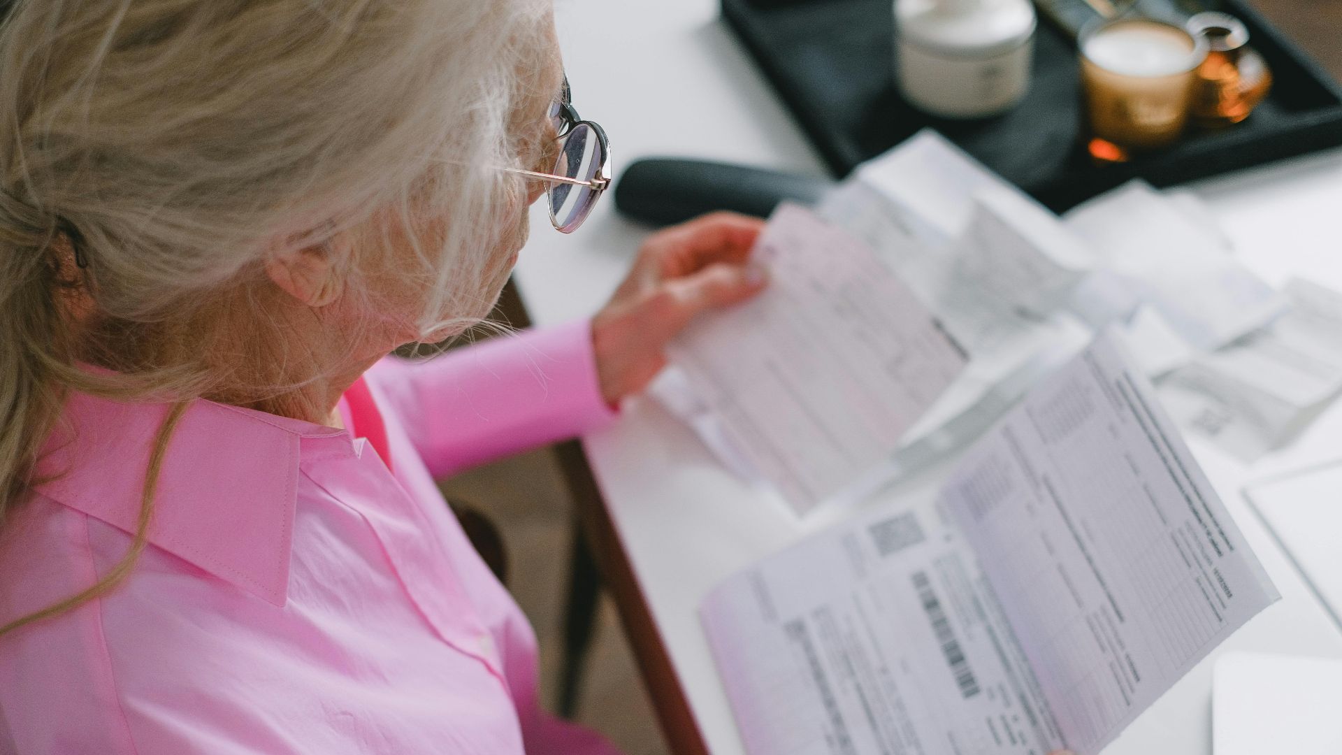 An elderly woman in glasses holds and reads important papers at a table indoors.