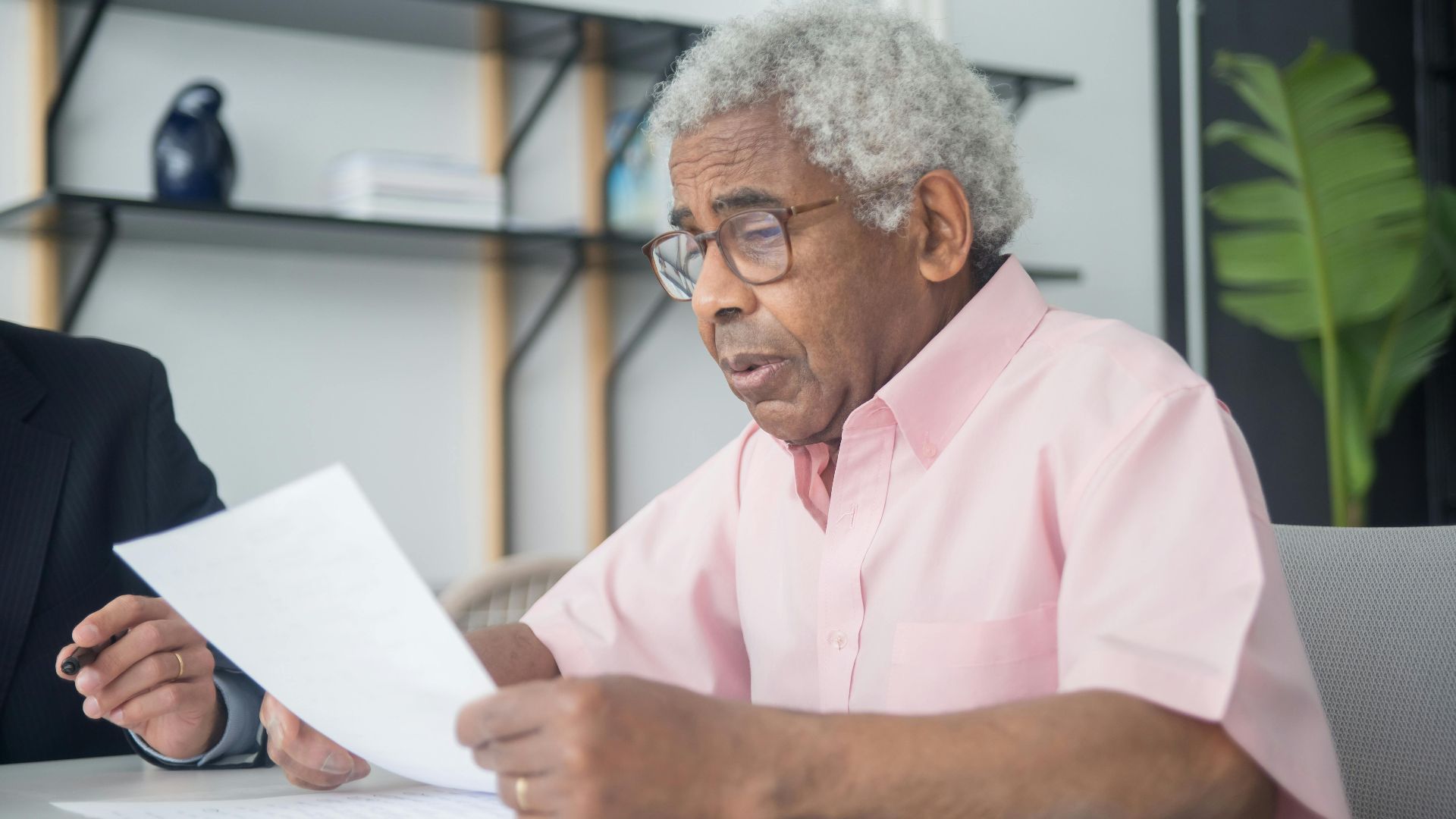 Elderly man with glasses reading a document in a home office setting, reflecting focus and concentration.