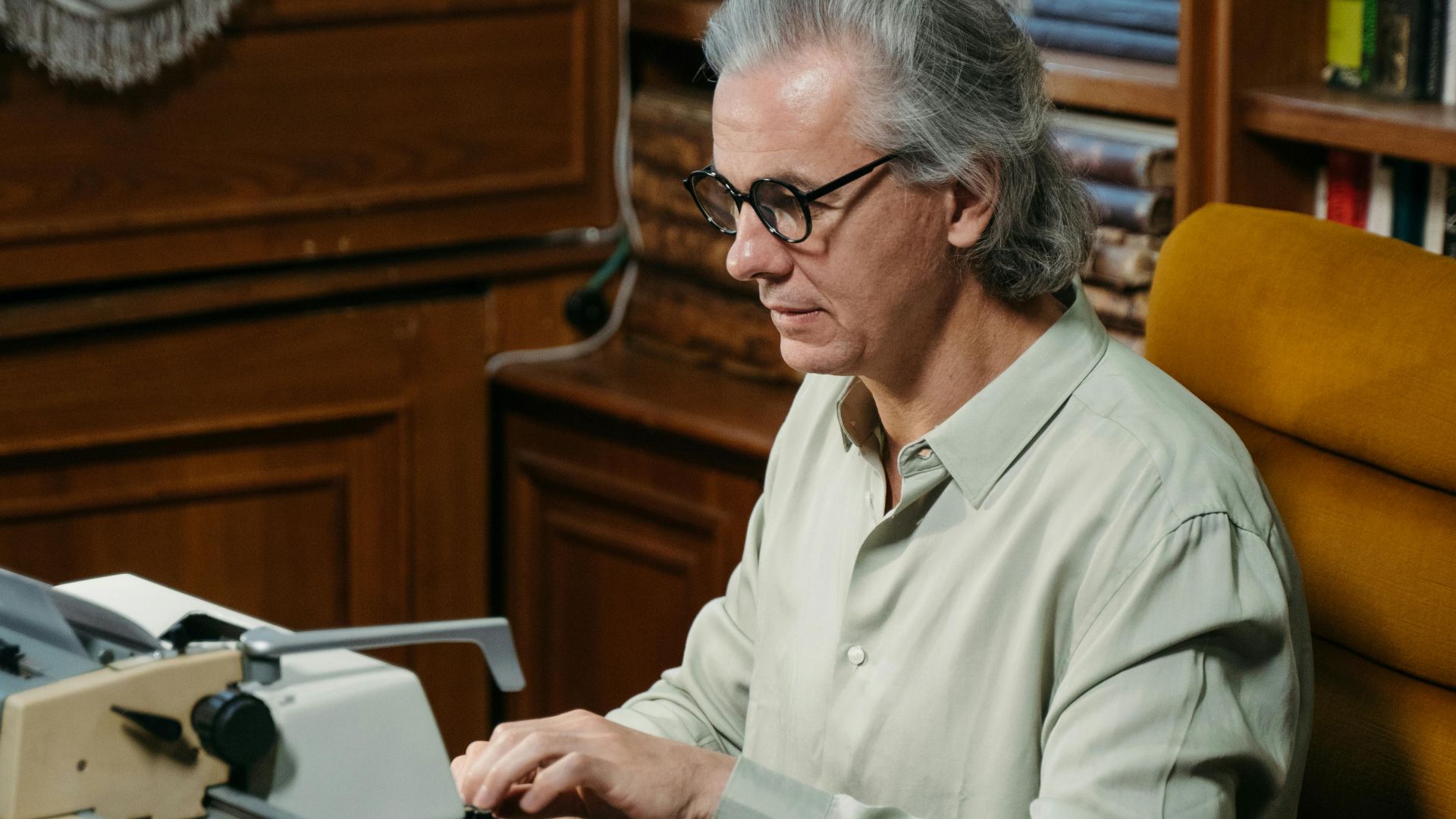 A senior adult man typing on a vintage typewriter in a classic library setting.