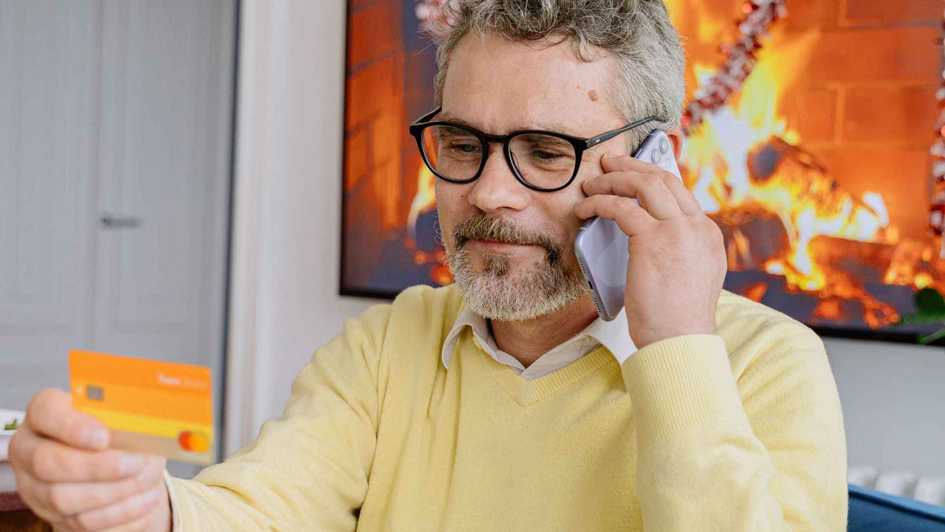Elderly man in yellow sweater using phone and credit card while sitting indoors by a fireplace.