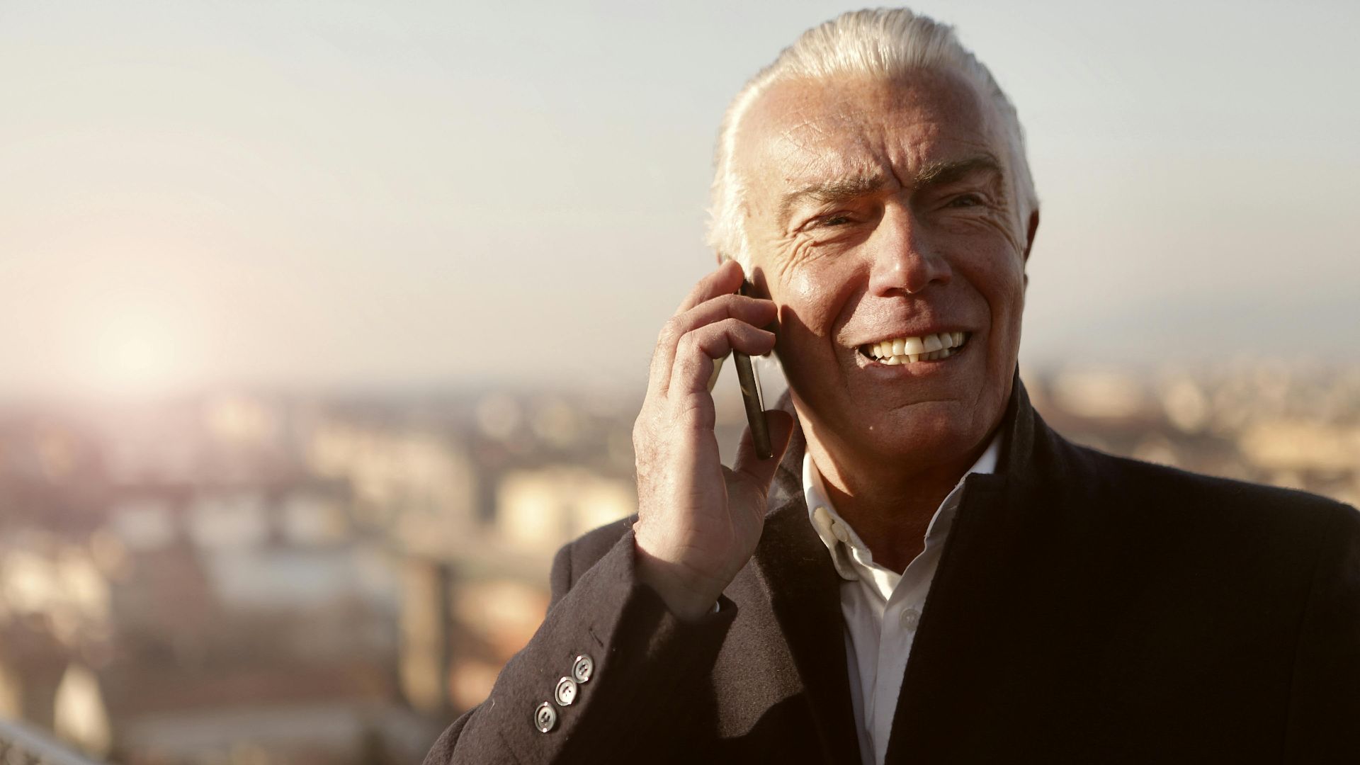 Senior man smiling as he talks on his smartphone with a cityscape background and setting sun.
