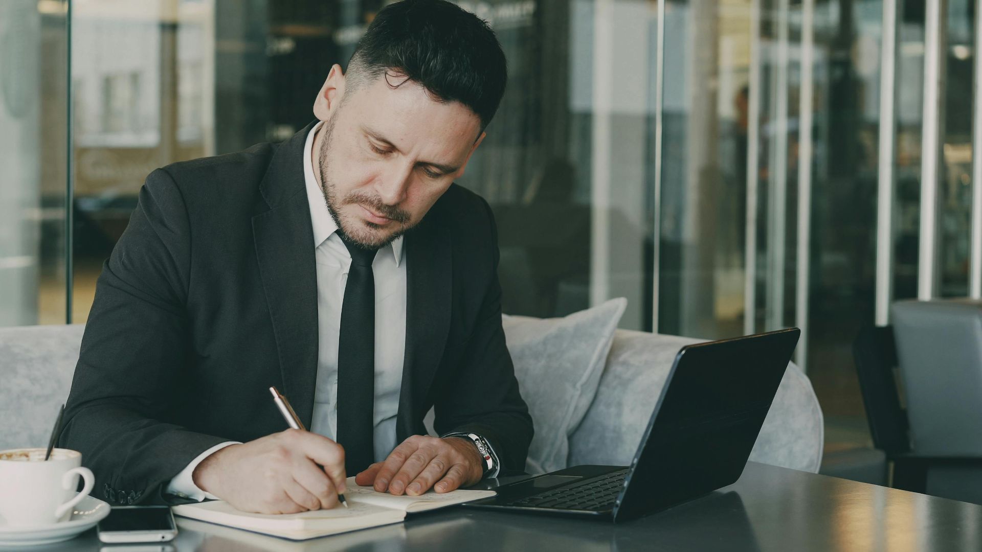 Professional businessman writing notes in a modern cafe setting, working on his laptop.