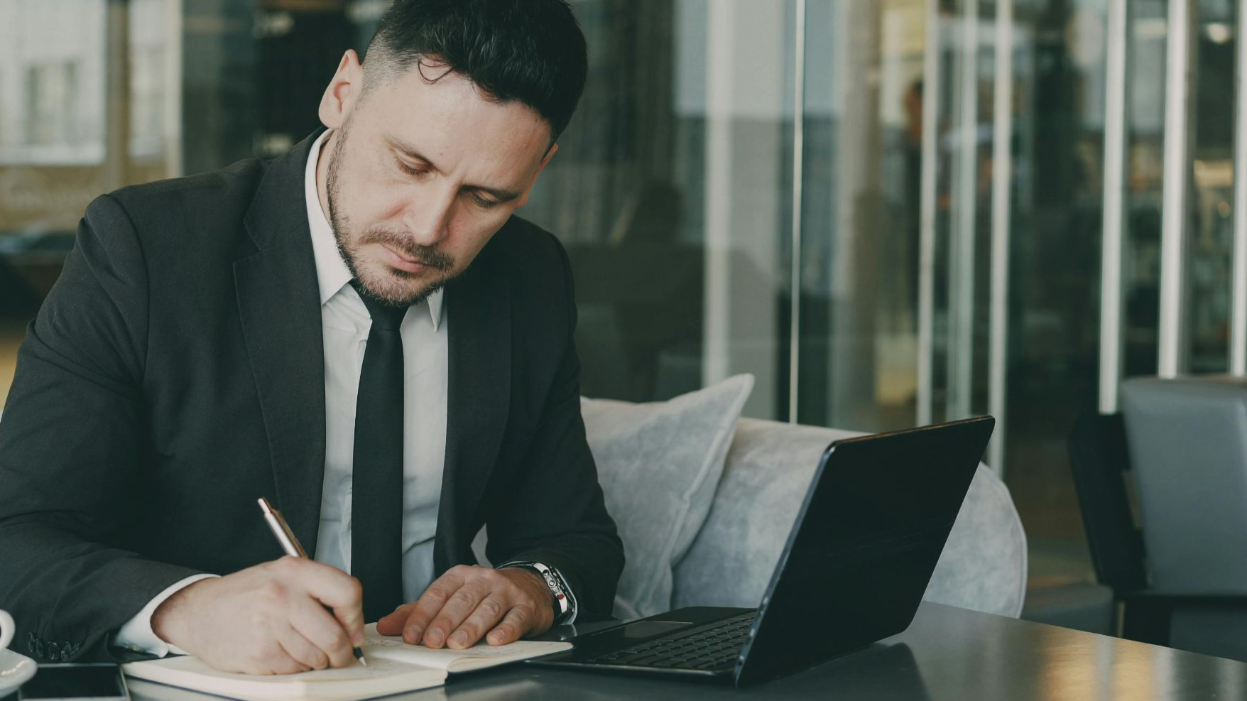 Professional businessman writing notes in a modern cafe setting, working on his laptop.