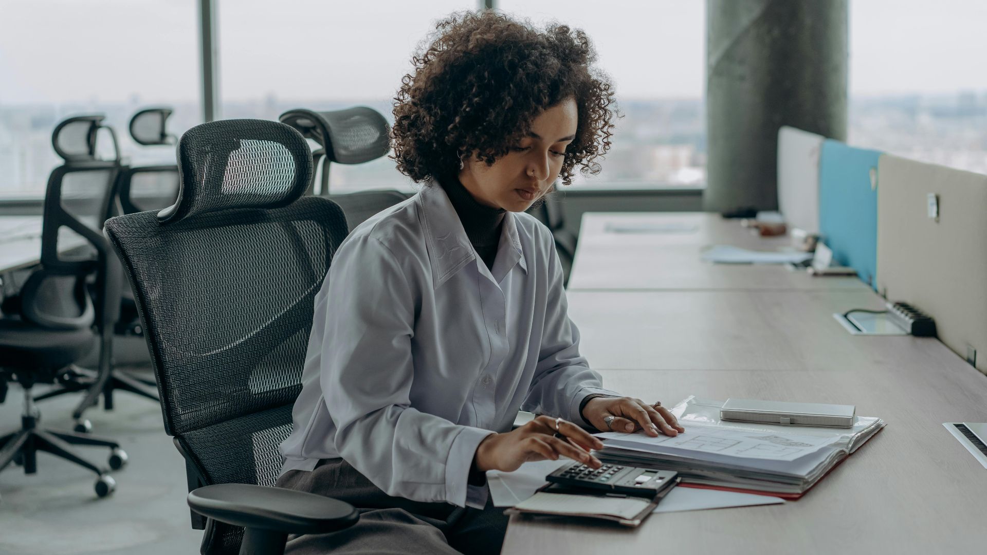 A professional woman with curly hair calculating documents in a modern office setting.