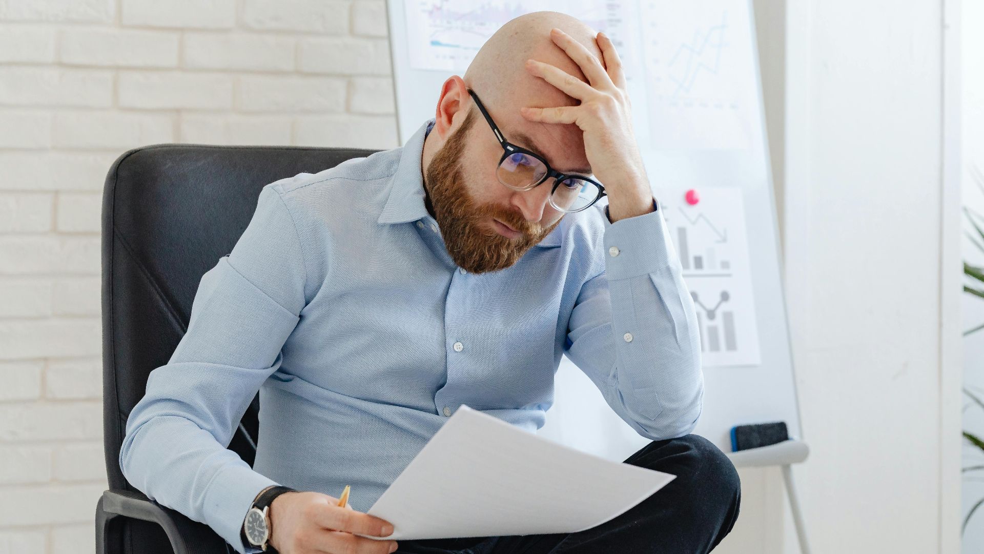 Bald man with glasses sitting in office looking worried at document.