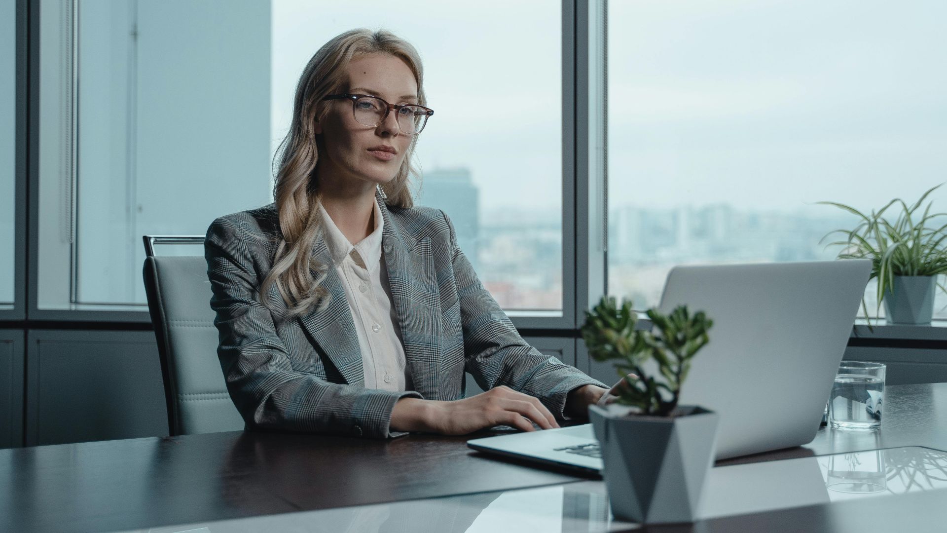 A confident businesswoman in a grey suit working on a laptop in a modern office setting.