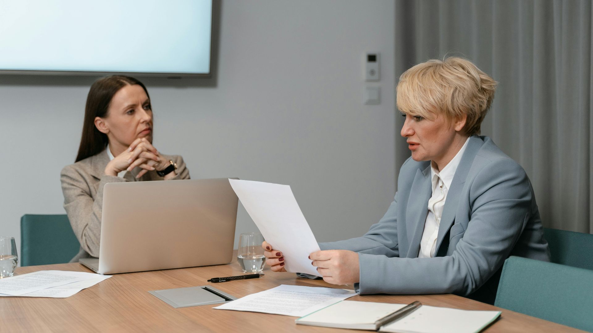 Two businesswomen in a serious meeting, discussing documents with a laptop in an office setting.