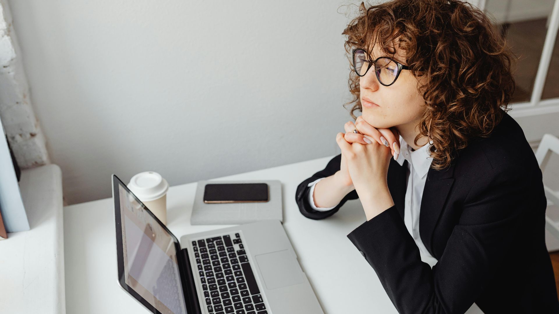 A pensive woman in business attire sits at a desk with a laptop, contemplating work.