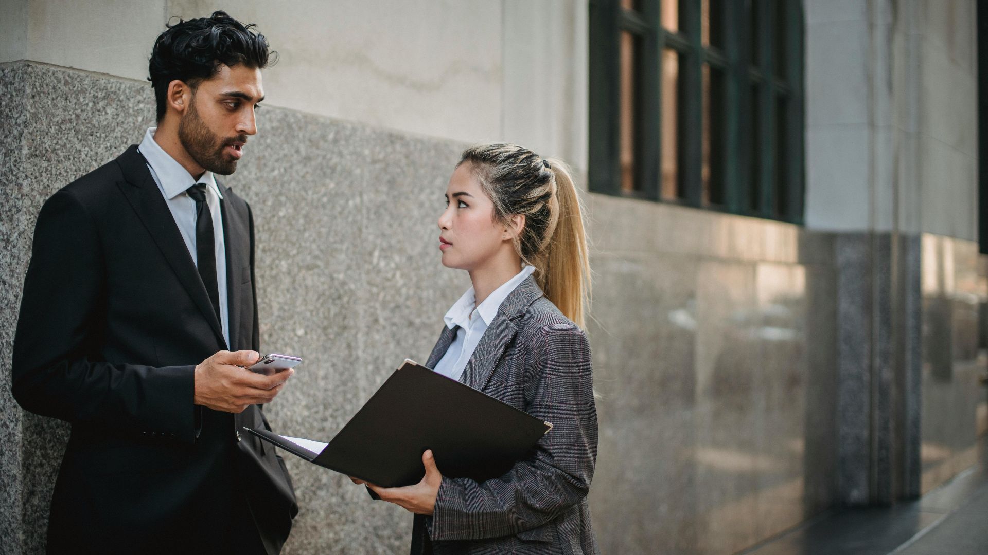 Two professionals in business attire discussing work outside an office building.