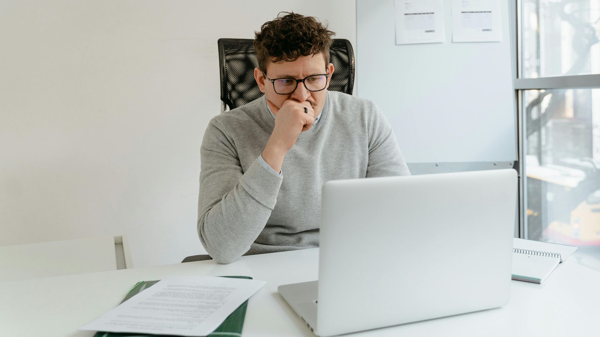 A thoughtful man in a gray sweater concentrates on his laptop at a bright office desk.