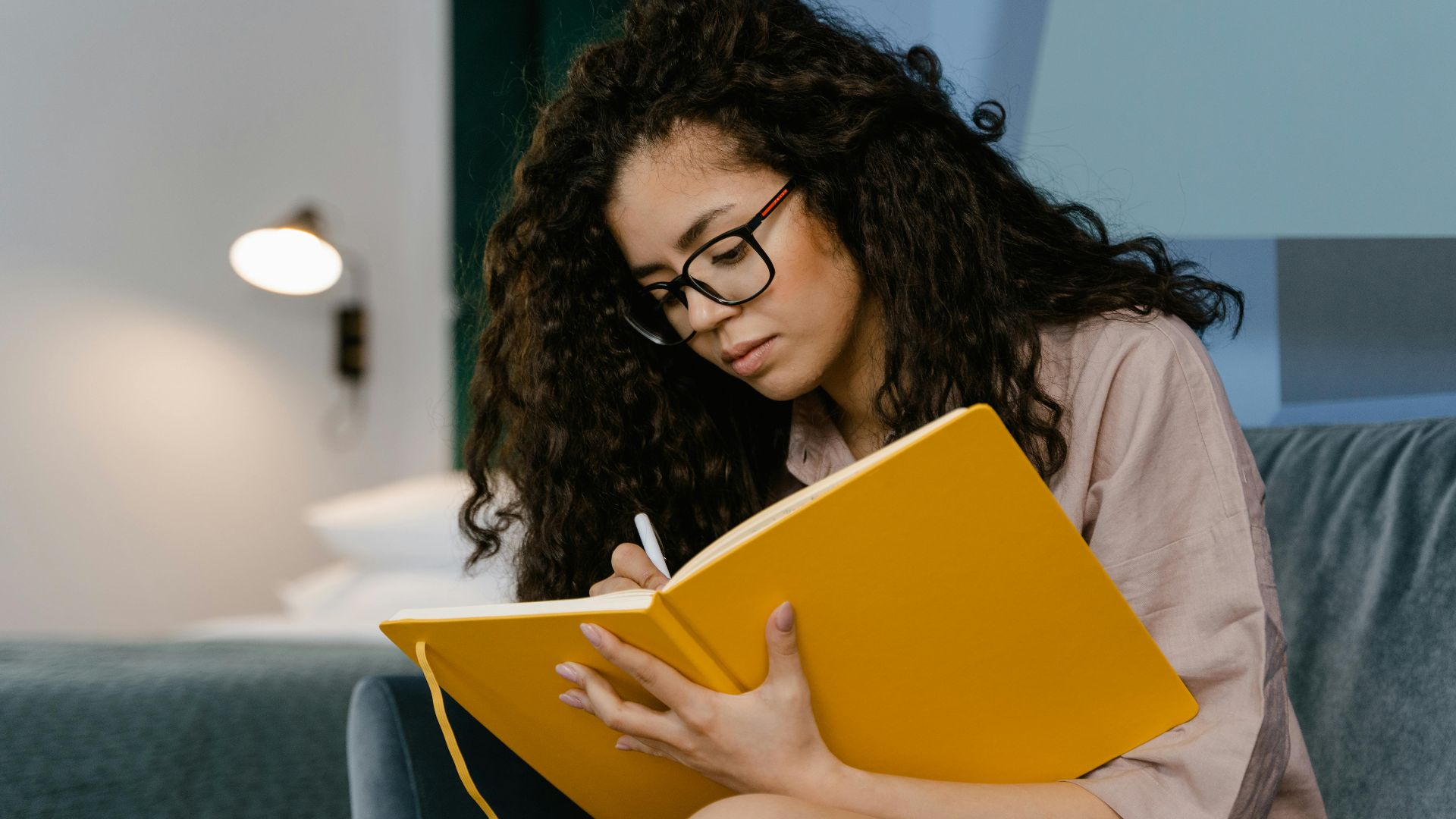 Young woman with curly hair writes in a yellow notebook, seated indoors.