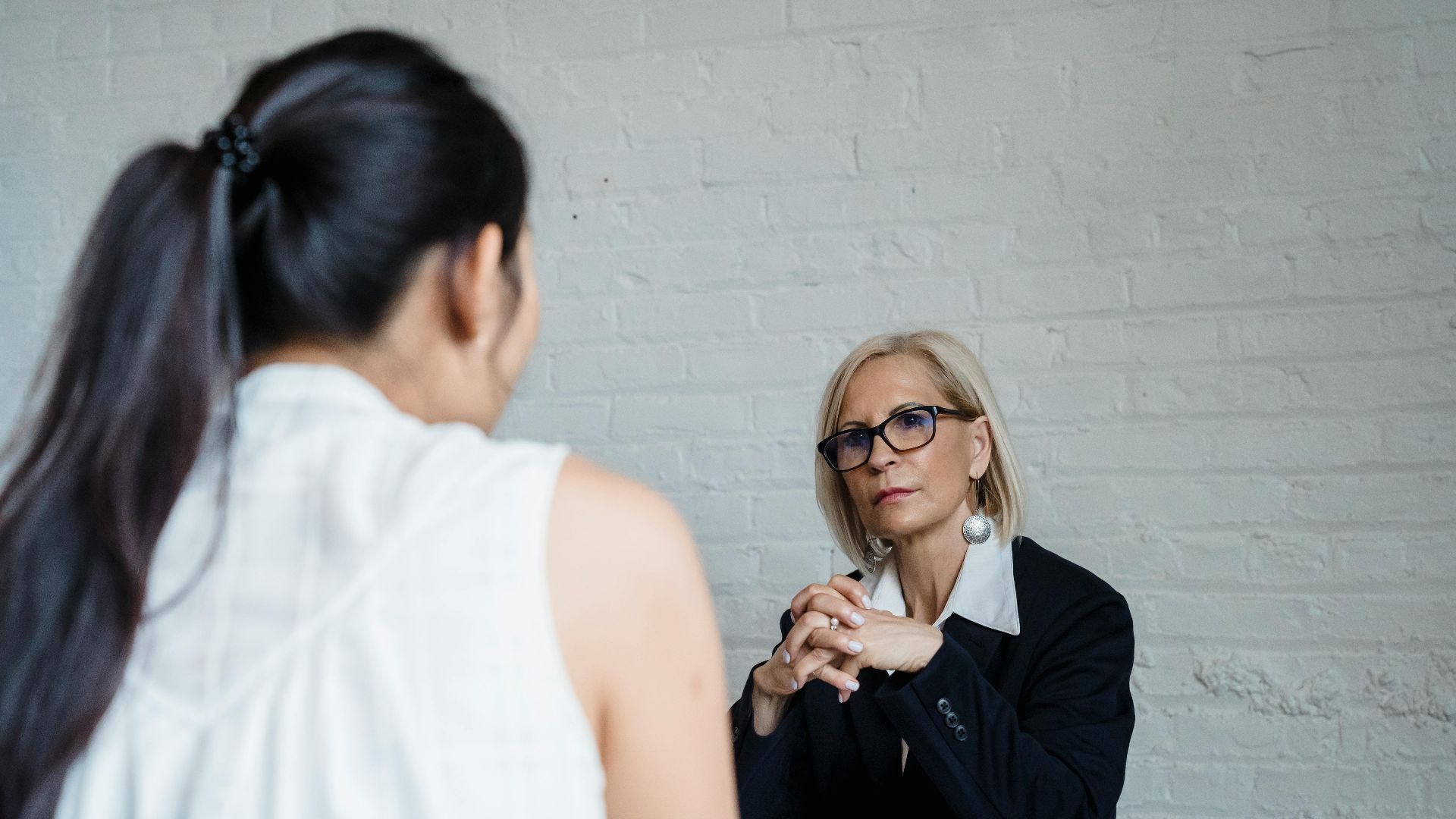 Two women having a serious conversation in an office setting against a brick wall background.