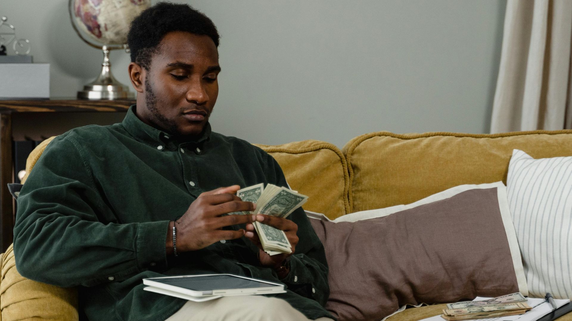 A man sitting on a couch counting cash notes in a cozy indoor setting with a globe in the background.