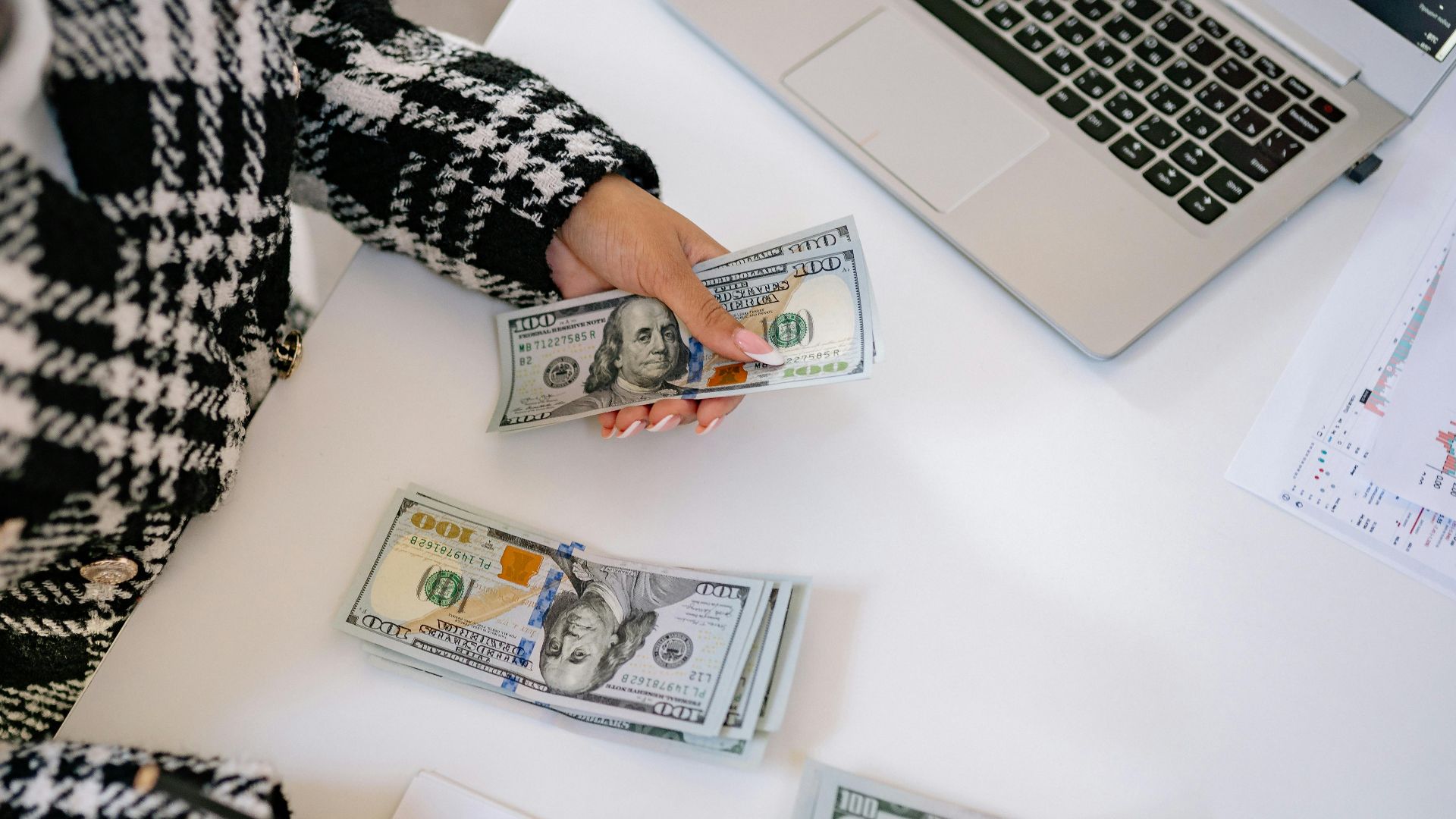A woman in a plaid coat counts American dollars at a desk with a notebook and laptop.