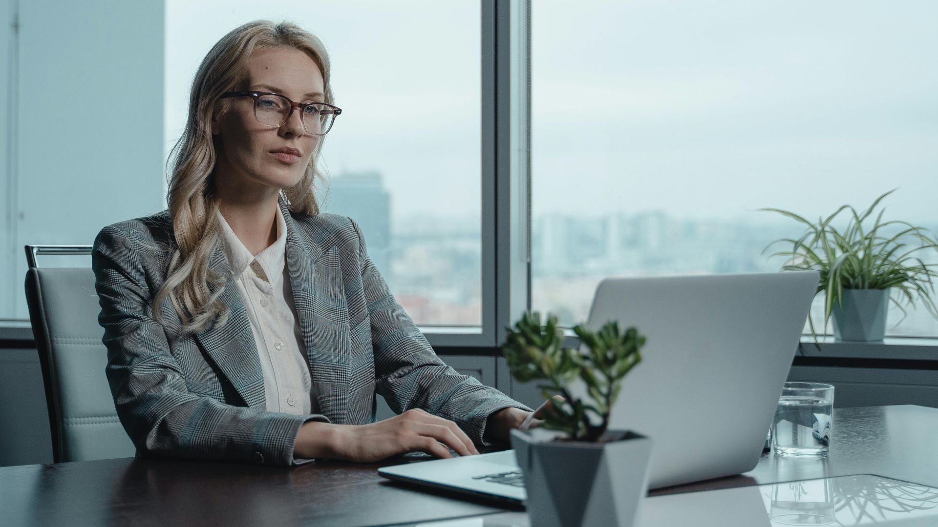 A confident businesswoman in a grey suit working on a laptop in a modern office setting.