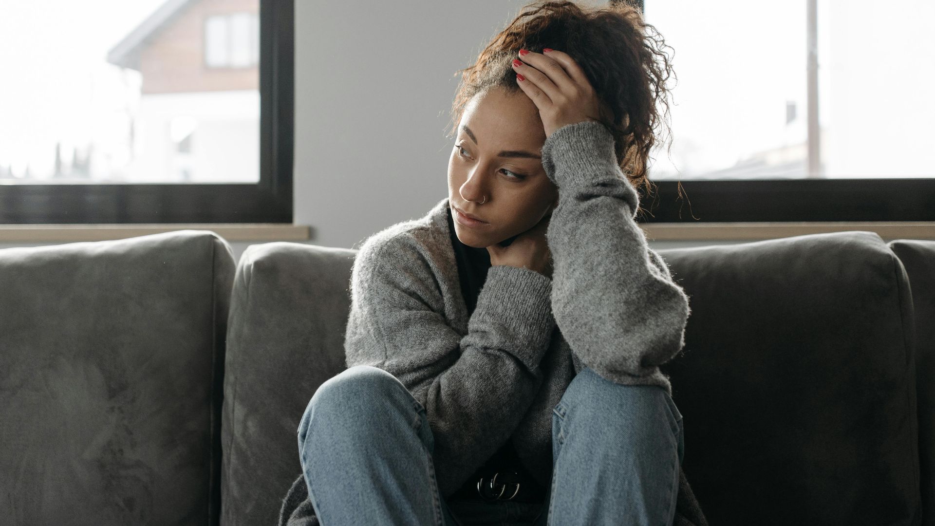 Thoughtful woman in cozy sweater sitting on sofa indoors by the window.