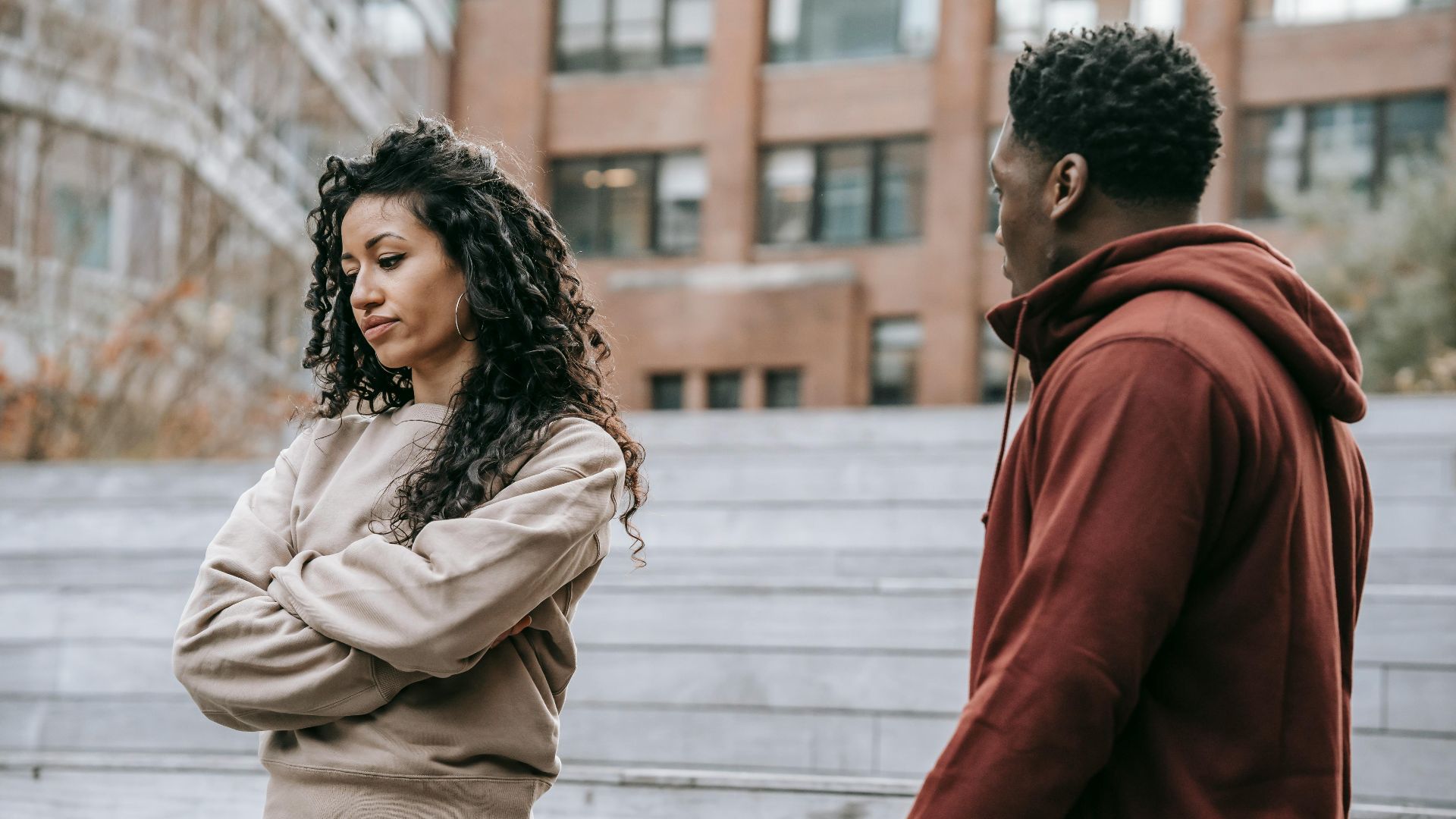 Offended ethnic girlfriend with curly hair standing with crossed arms standing near African American boyfriend while having conflict on street in park