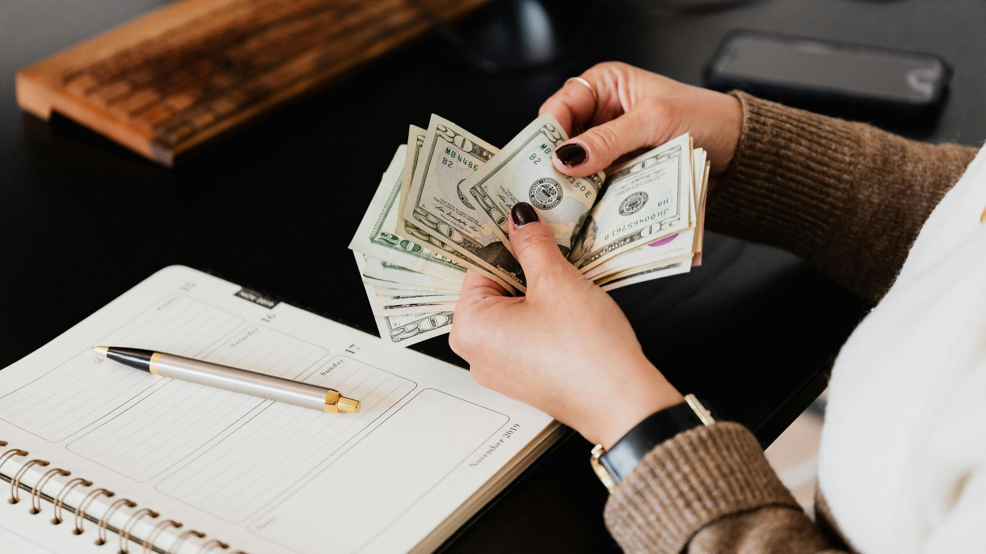 Unrecognizable elegant female in sweater counting dollar bills while sitting at wooden table with planner and pen