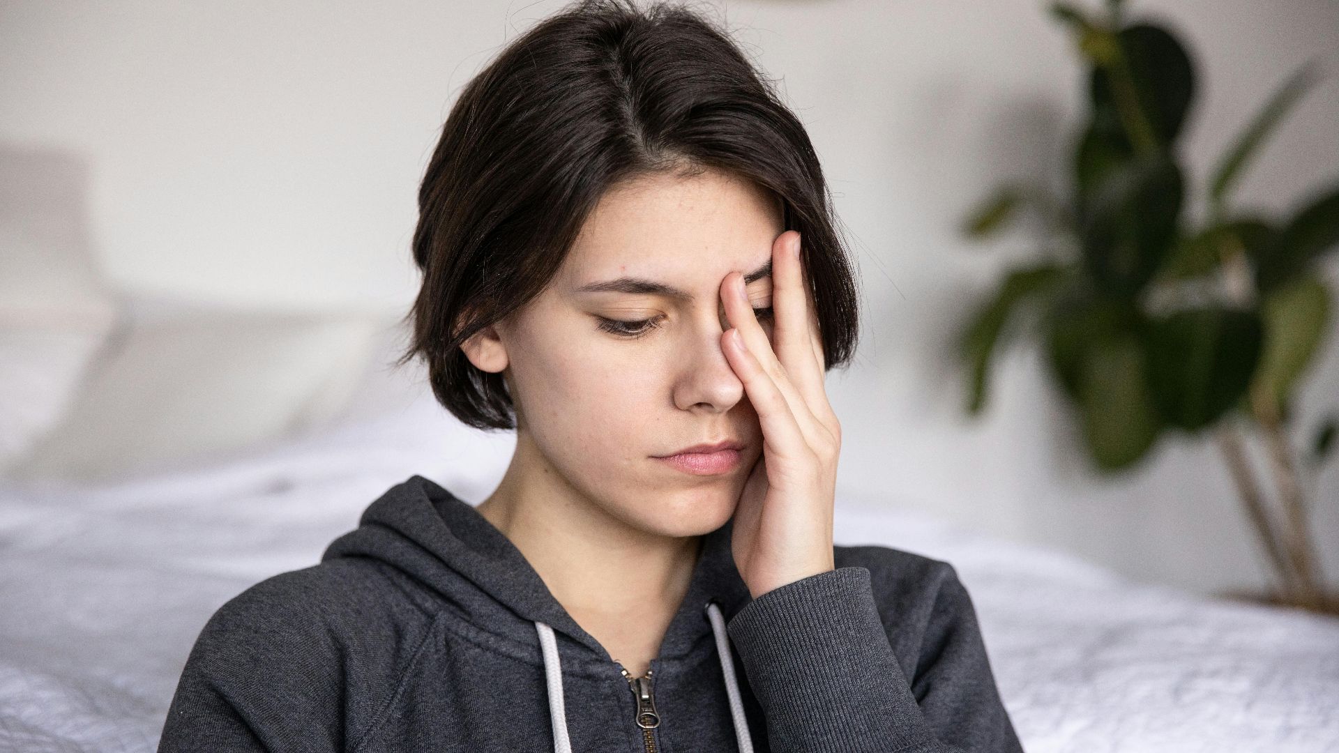 A young woman in a grey hoodie looks contemplative while sitting indoors, hand resting on her face.