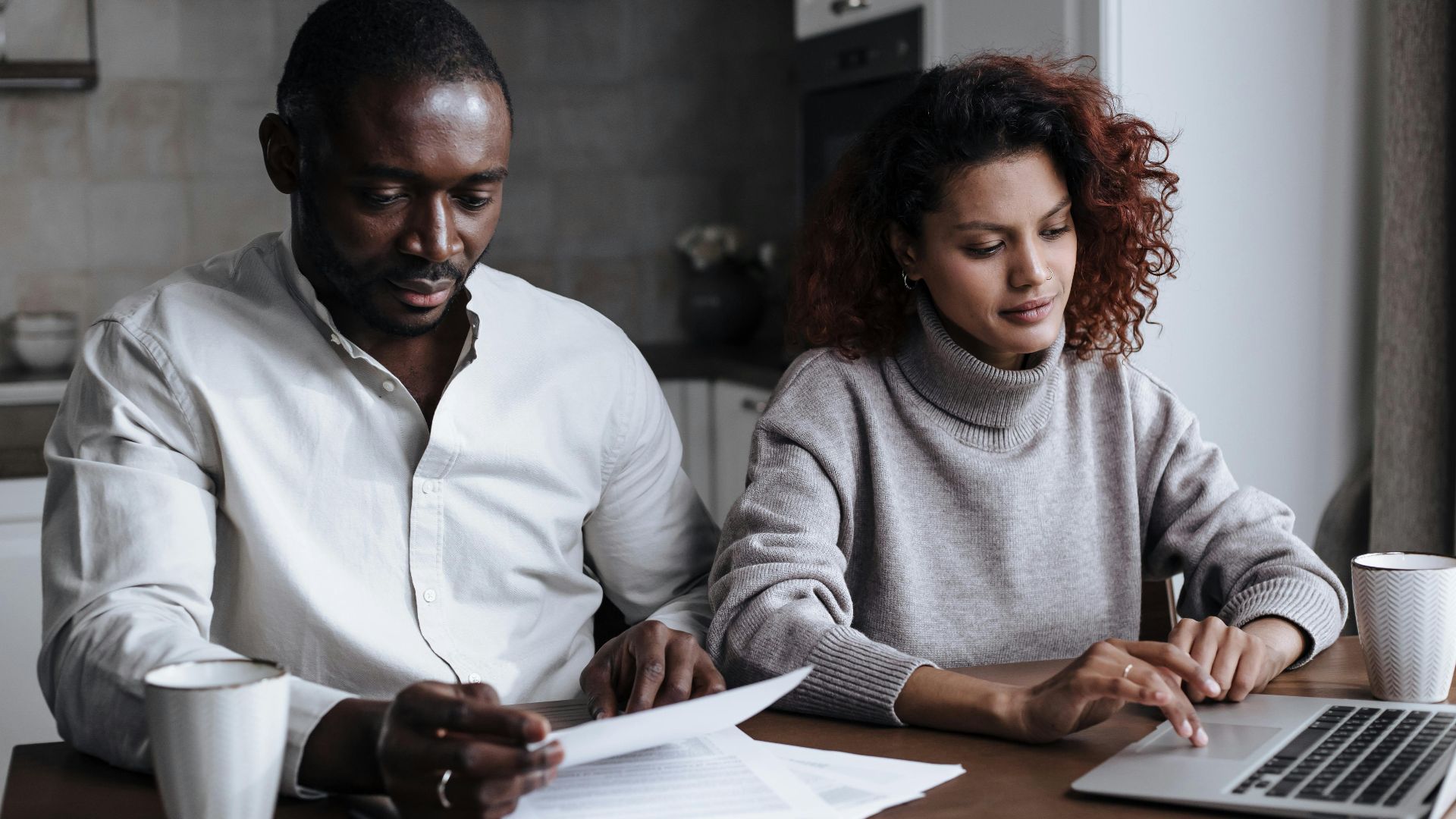 A diverse couple collaborates on a laptop at their home, handling documents over coffee.