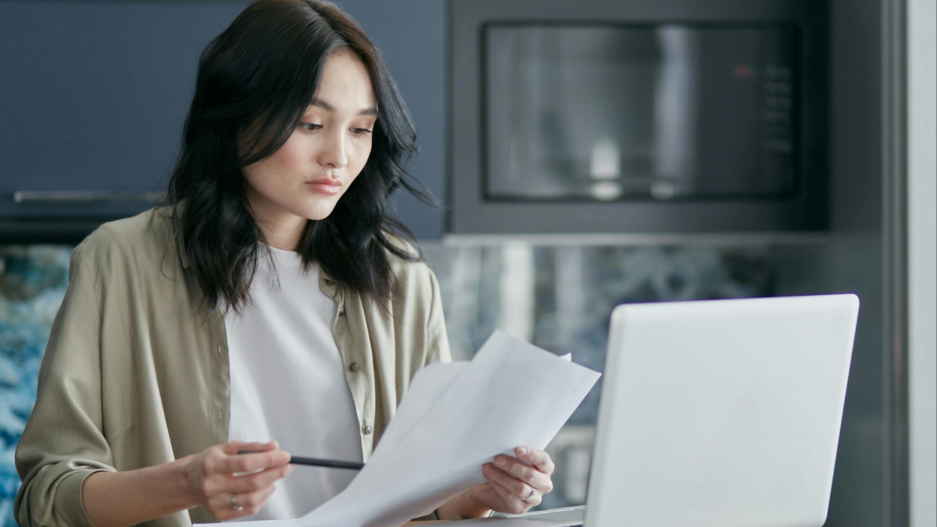 Concentrated woman reviewing documents and working on a laptop at home office setup in kitchen.