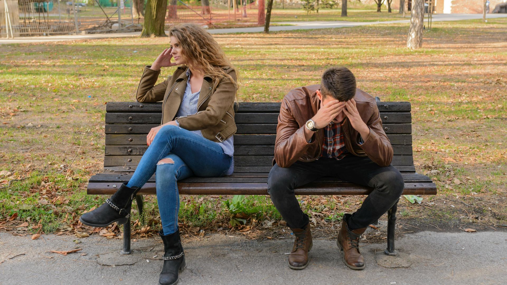 An upset couple seated on a park bench, expressing frustration during an autumn day.