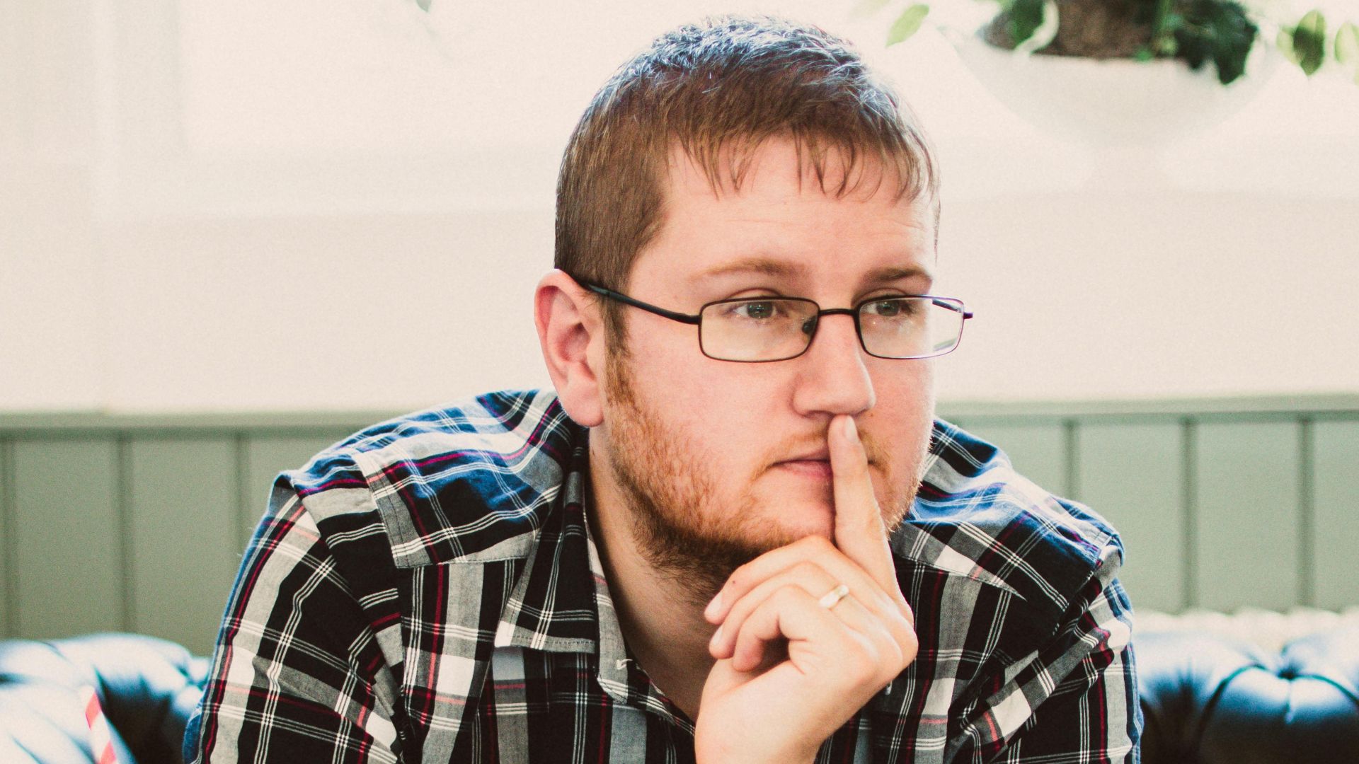Young man wearing glasses thinking while writing in a cafe with a drink beside him.