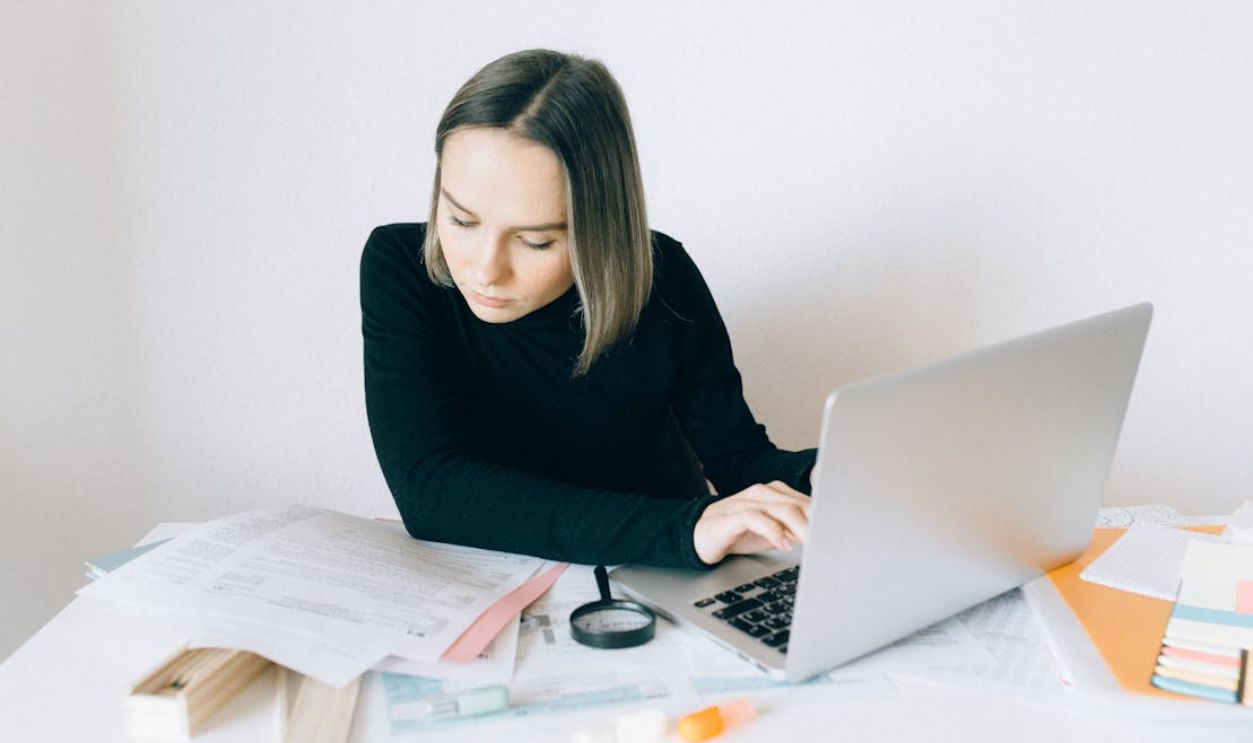 A Woman in Black Long Sleeves Doing Paperwork while Typing on Her Laptop