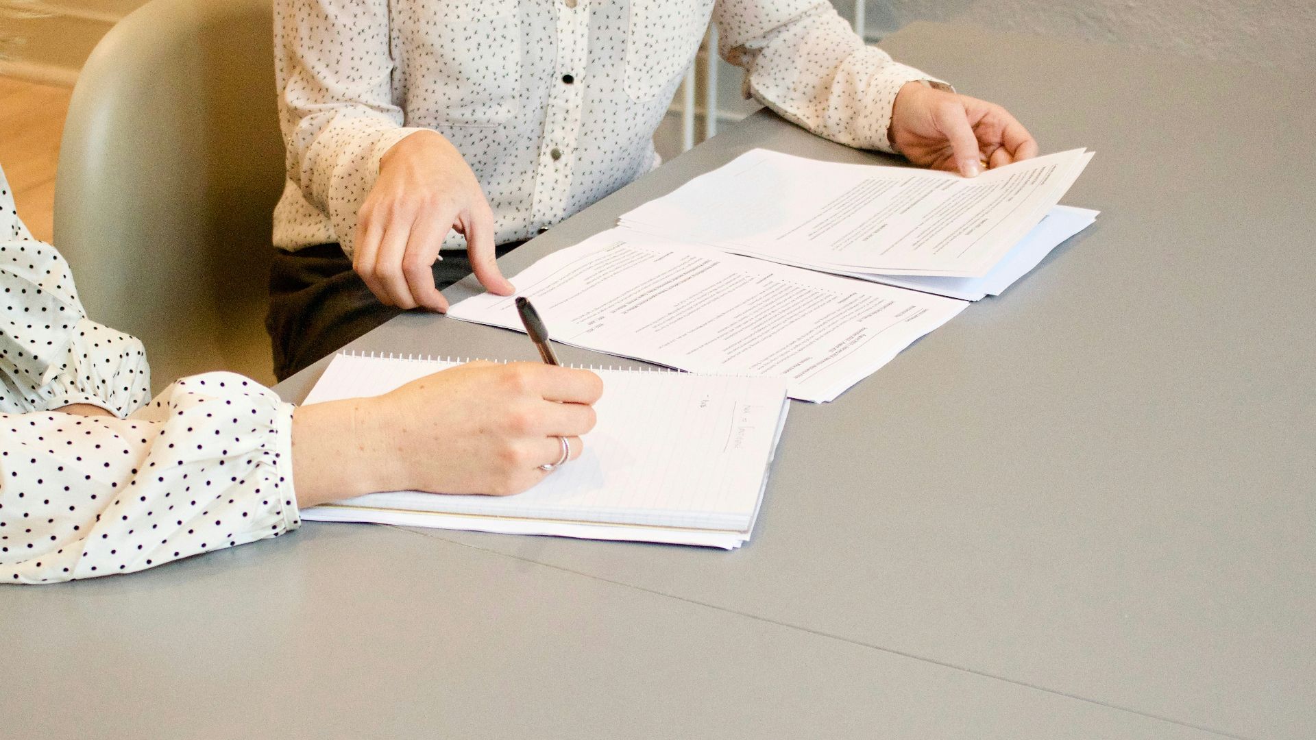 woman signing on white printer paper beside woman about to touch the documents
