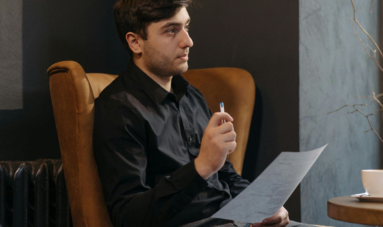 Man in Black Long Sleeve Shirt Sitting on a Chair While Holding a Paper