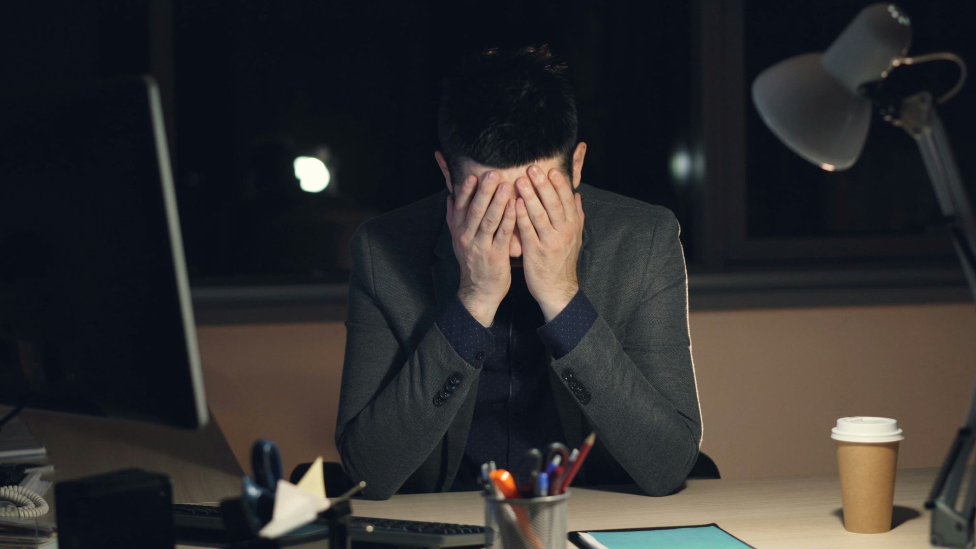 Man in suit sits at desk, head in hands.