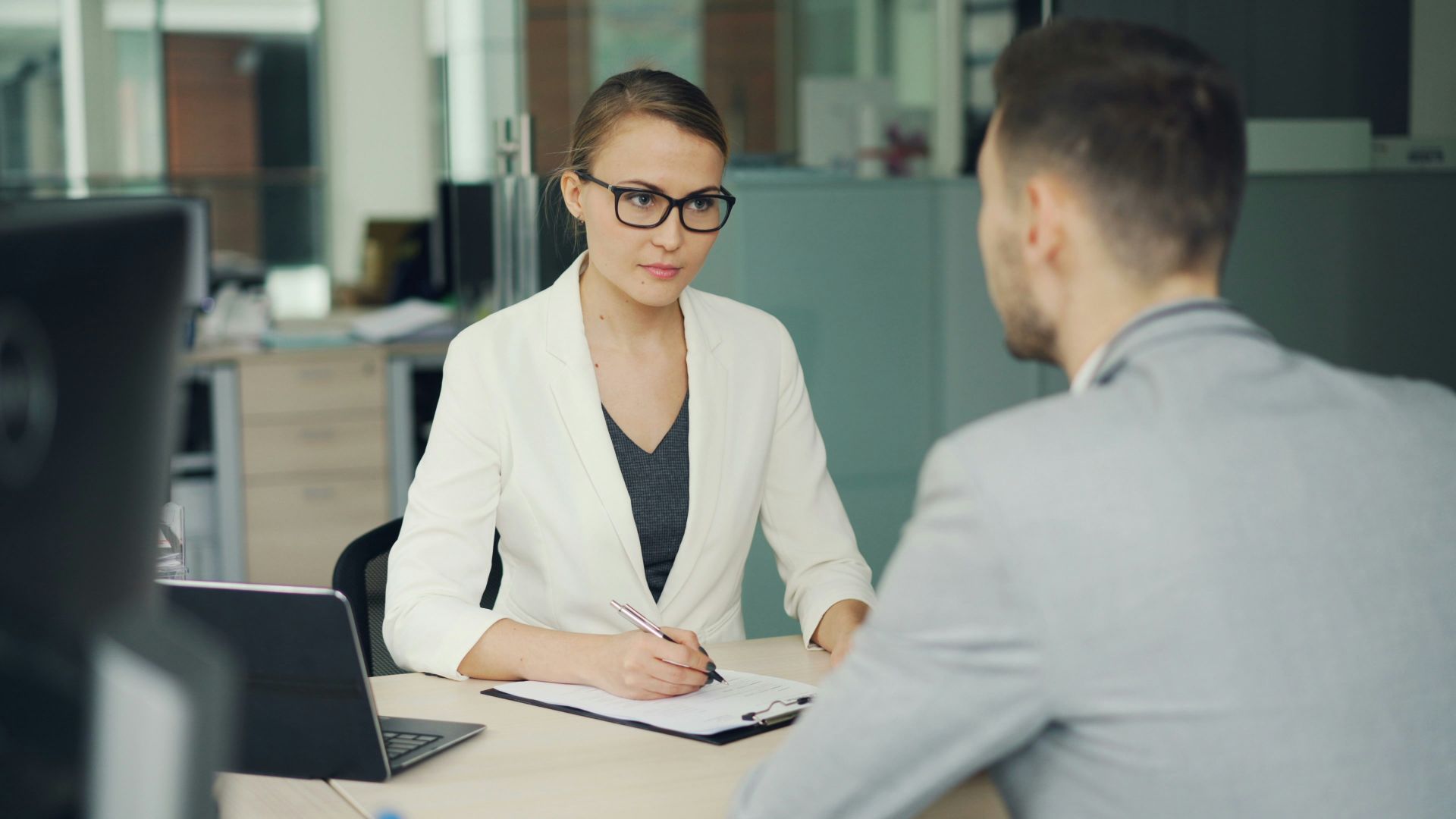 Woman in glasses interviews man at office desk.
