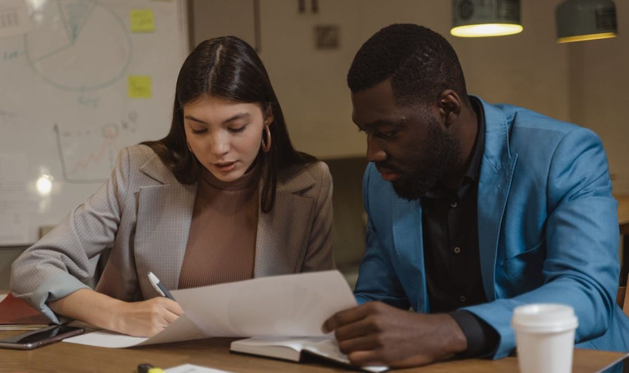 A Man in Blue Suit Jacket Beside Woman in Gray Blazer Writing on Paper